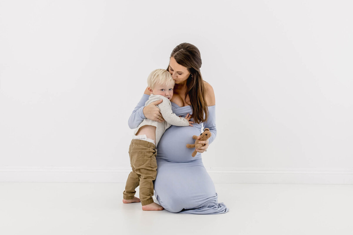 A pregnant woman in a light blue dress kneels on a white floor while hugging a young child with blond hair. The child, wearing a gray long-sleeve shirt and brown pants, holds a small stuffed animal.