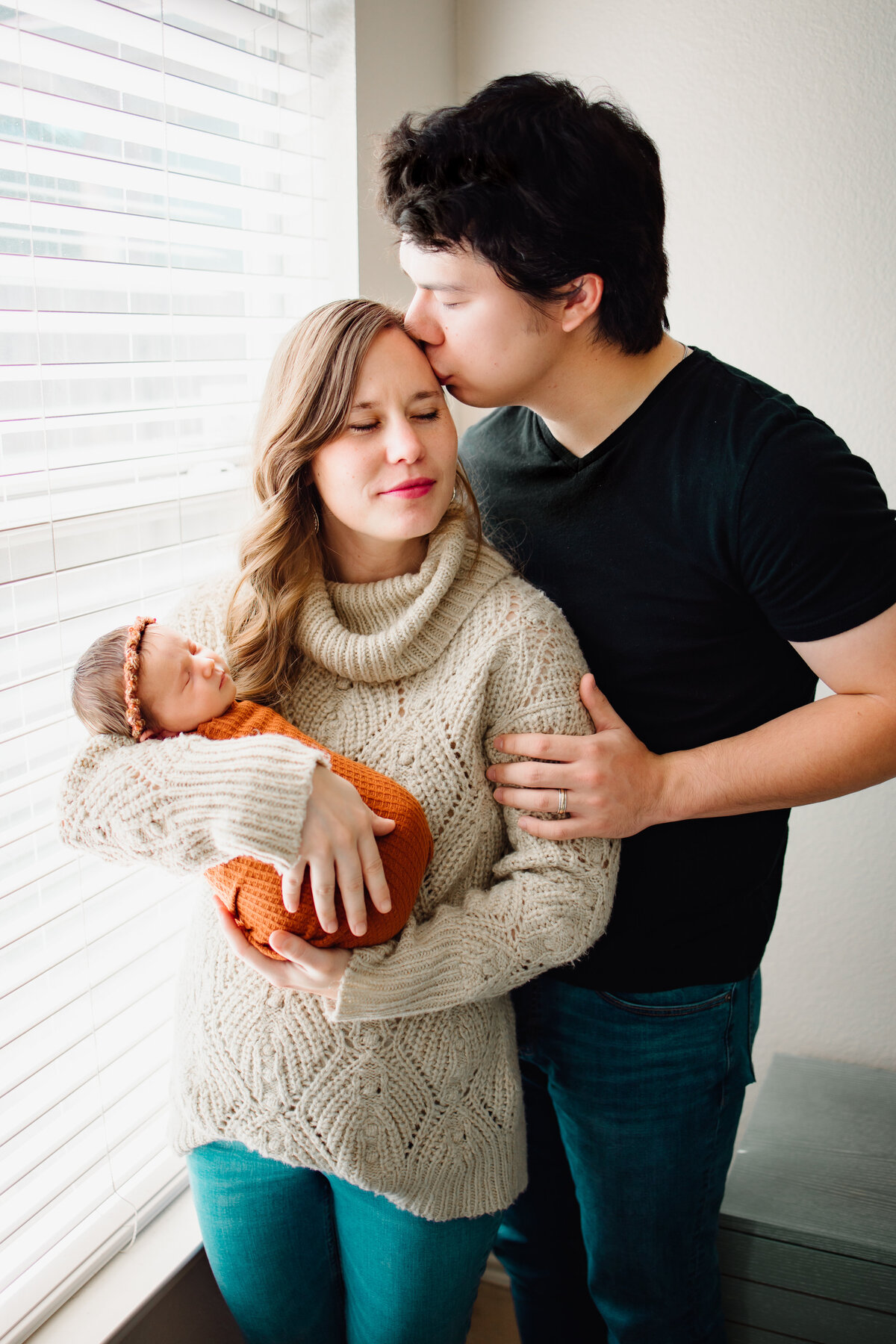 An affordable maternity photographer in Albuquerque captured this incredible family moment. The beautiful mommy is hugging her newborn baby wrapped in a cozy orange blanket, while the dad lovingly kisses her face.