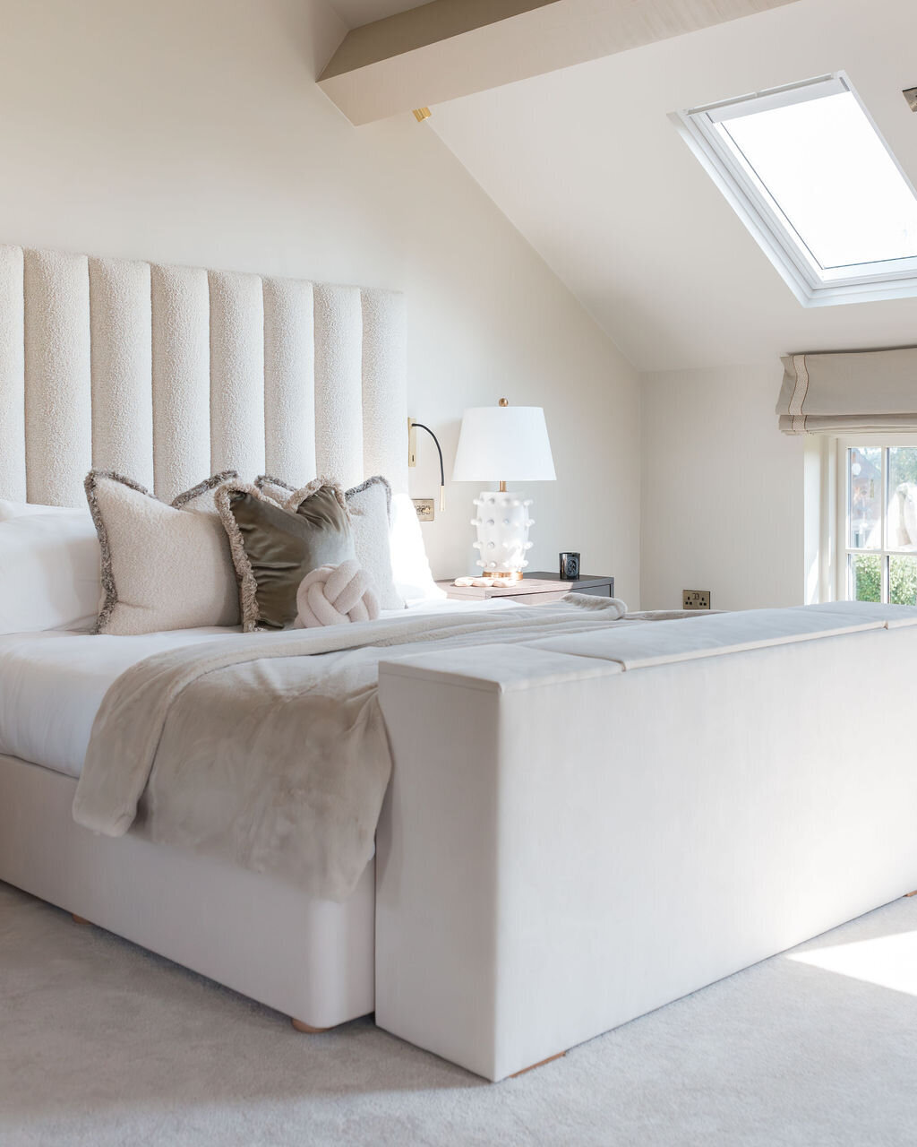 A well-lit bedroom featuring a large plush bed with a tall, tufted headboard. The room has a neutral color palette with white and beige bedding and some accent pillows. A skylight lets in soft natural light.