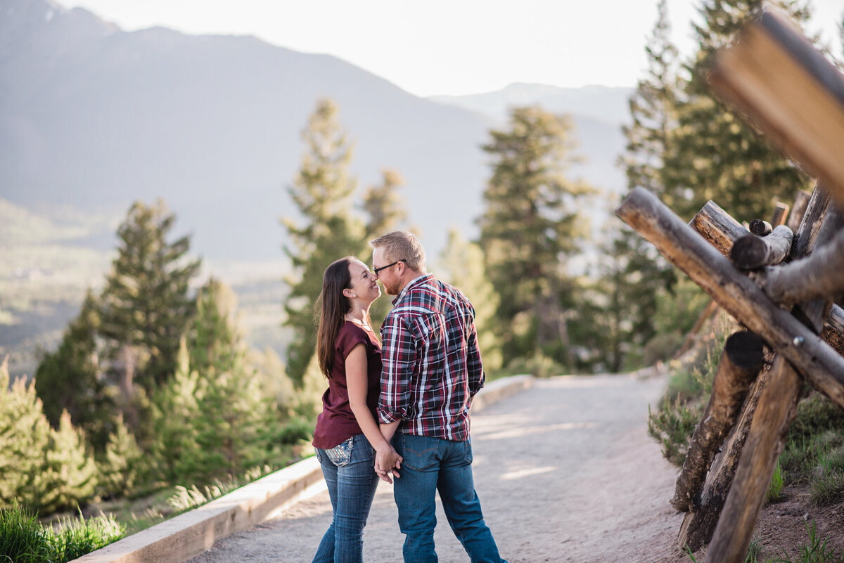denver engagement photographer captures outdoor engagement pictures with man and woman holding hands while walking down a back road as they lean in and kiss each other with the. mountains and woods in the distance