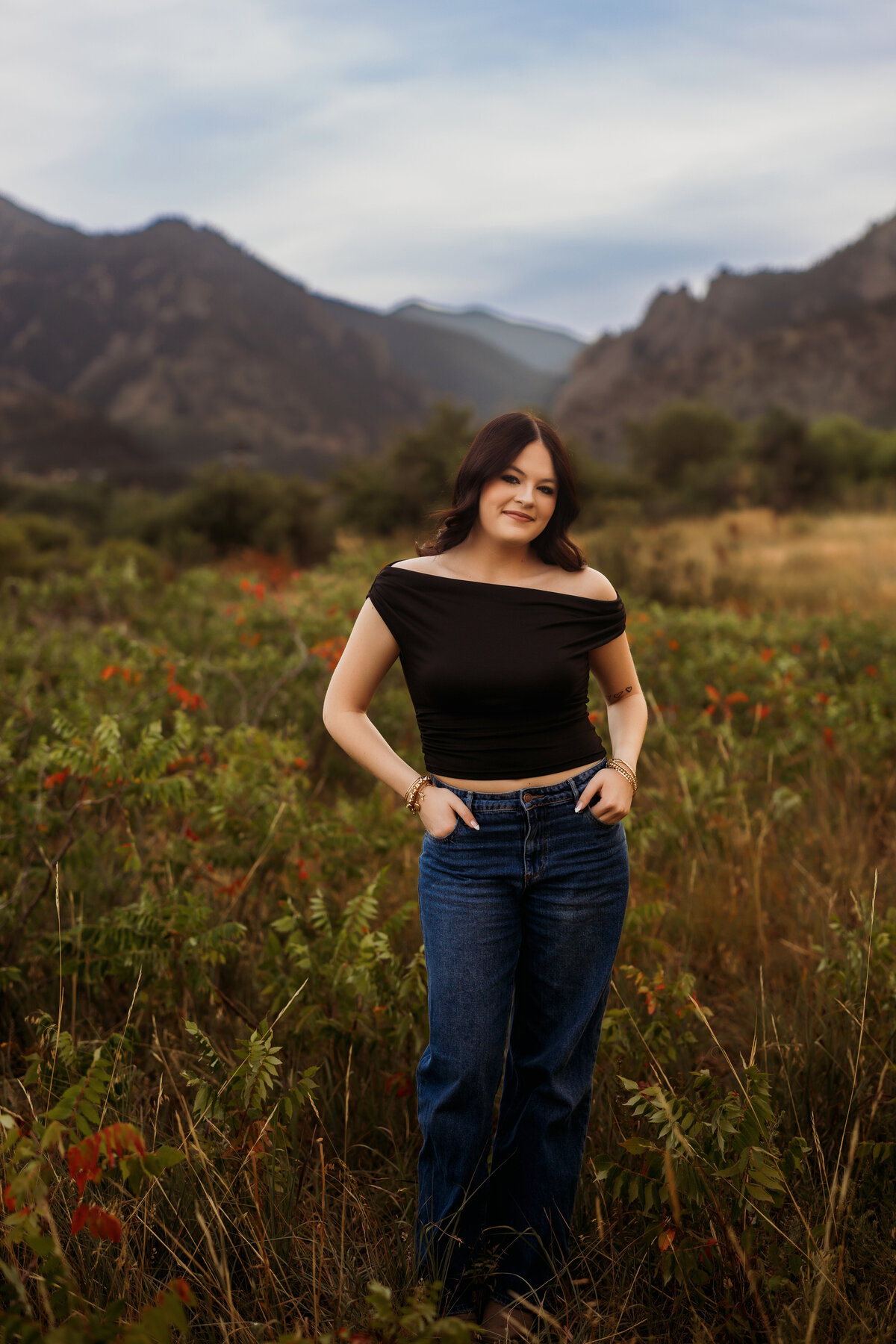 Senior girl stands in the middle of tall grasses and nature with mountains in the background for her senior photos just outside of Denver