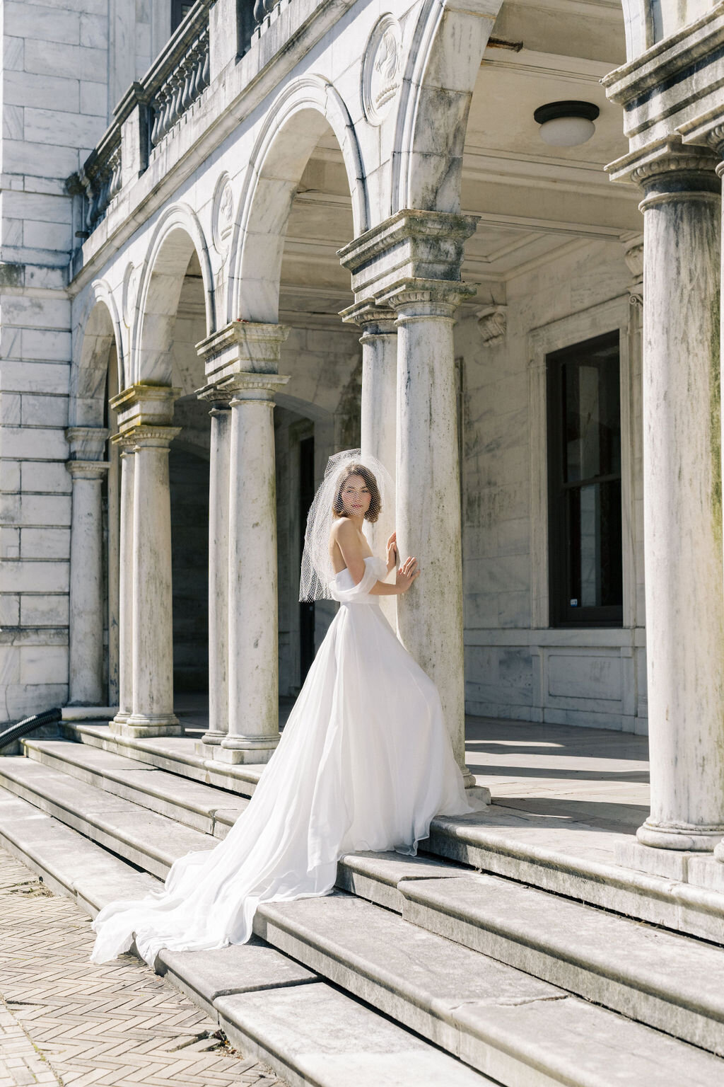 bride wearing wedding dress with long train leaning against mansion column