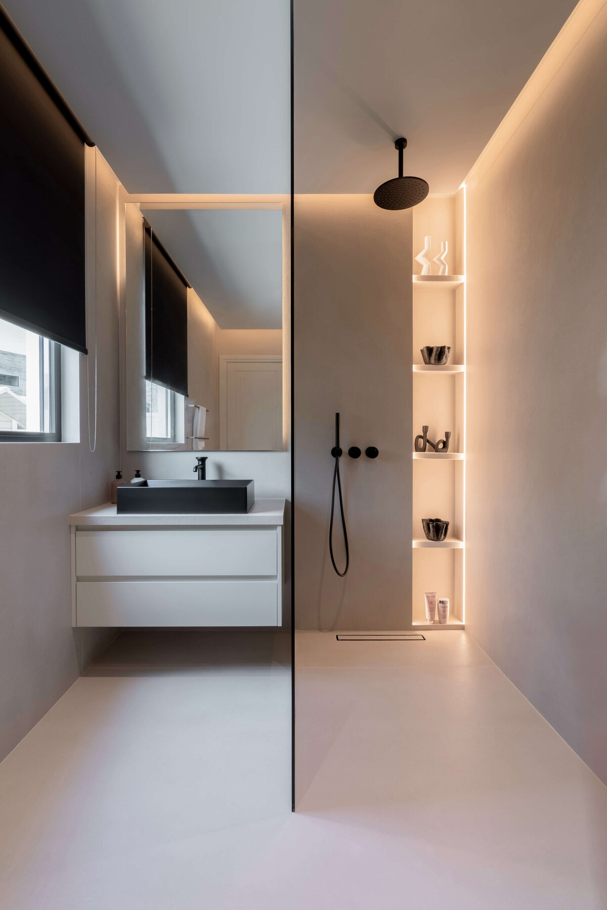 Modern, minimal bathroom with a sink on one side and a softly lit walk-in shower with black hardware on the right.