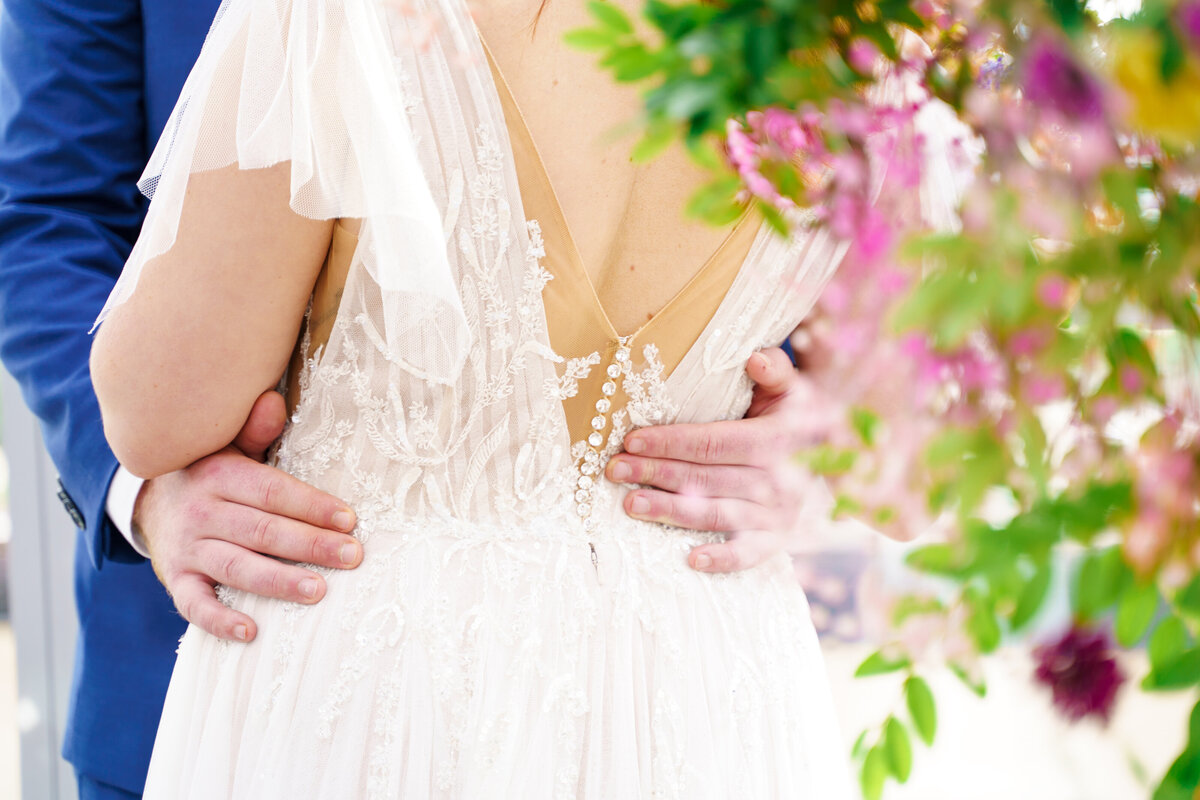 Groom holds his bride's waist and her dress details of beads and draped chiffon with flowers in the foreground. Wedding at the North Fourth Corridor (The Revery) in Columbus, Ohio.