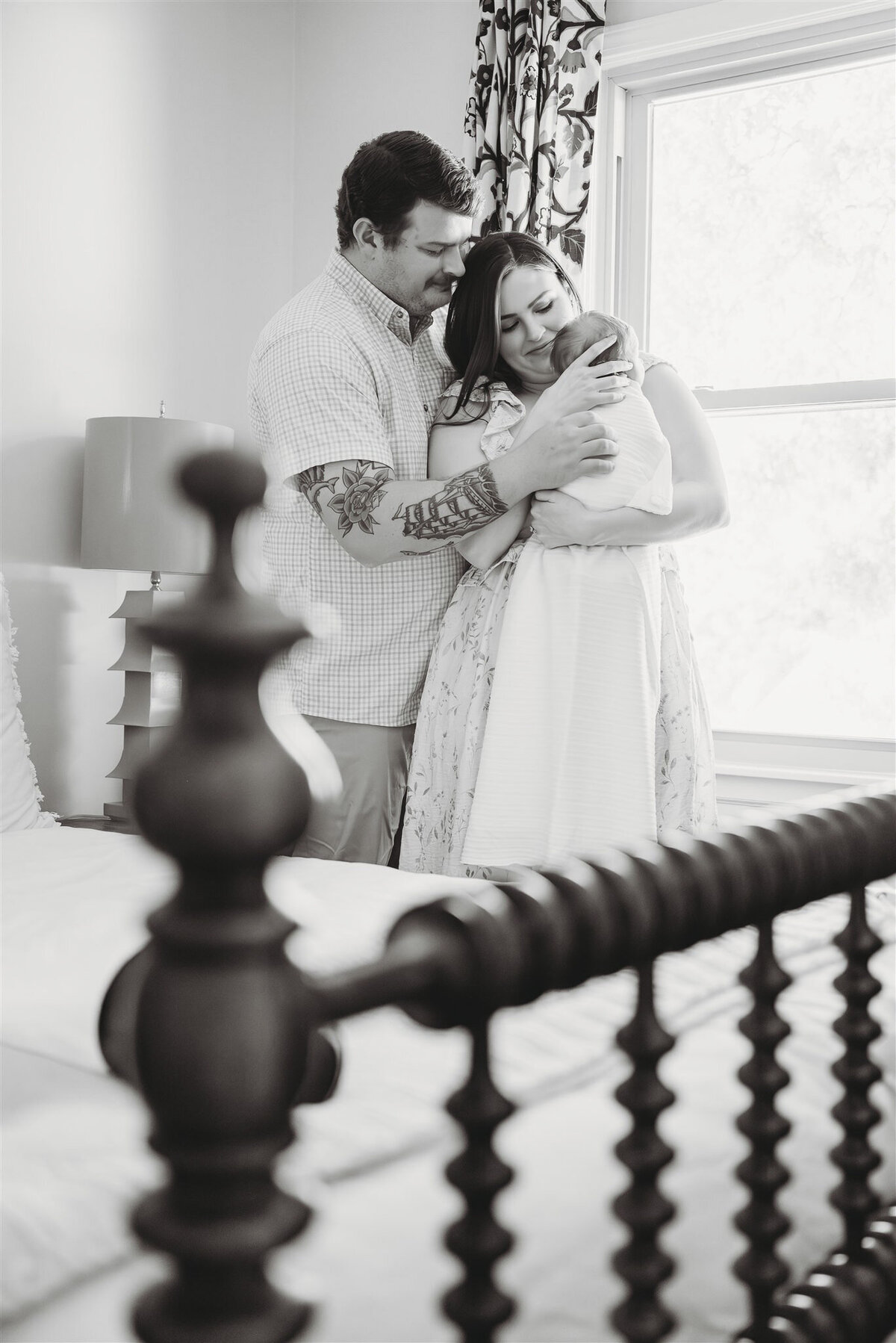 Black and white image of a mother holding her baby while standing near a crib, creating a timeless family portrait.