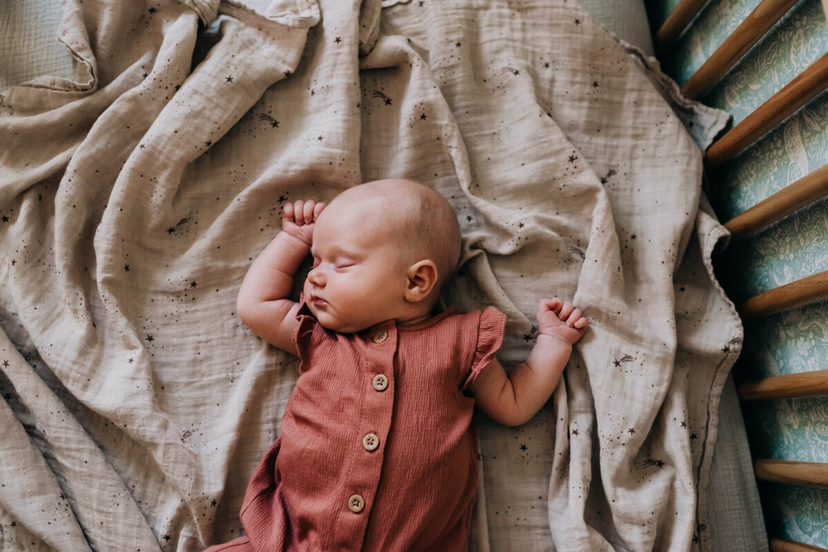 A baby dressed in a rose colored outfit sleeps on top of a tan swaddle in her crib