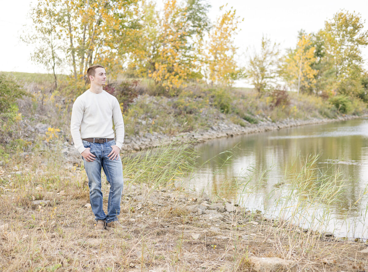 high school senior male, standing next to a pond, with trees in the background, thumbs in his pockets, wearing a light tan long sleeve shirt, blue jeans, a belt, and tan dress shoes, looking away from the camera towards the water
