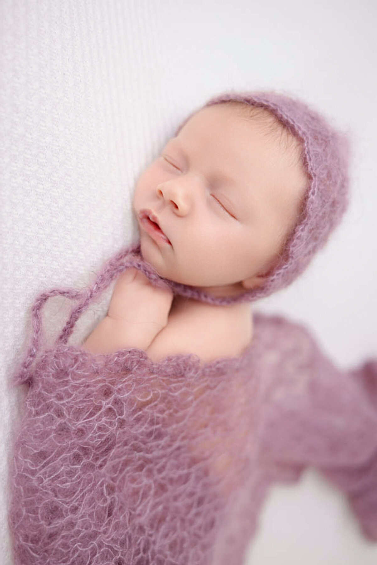 two-week baby girl posed lying on white blanket and wearing a purple bonnet and covered in a lacy purple baby wrap.