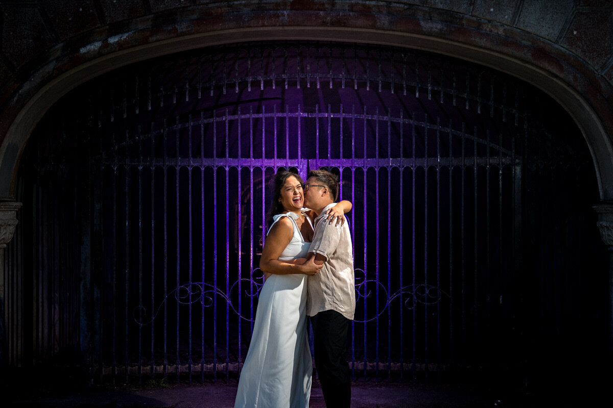 This playful and candid engagement photo captures a couple enjoying a lighthearted moment in front of an ornate iron gate across from the Conservatory of Flowers in San Francisco. The backdrop, enhanced with rich purple lighting, adds a moody and romantic touch to the image. The couple's laughter and joyful expressions stand out beautifully, creating a fun yet intimate vibe in this iconic setting.