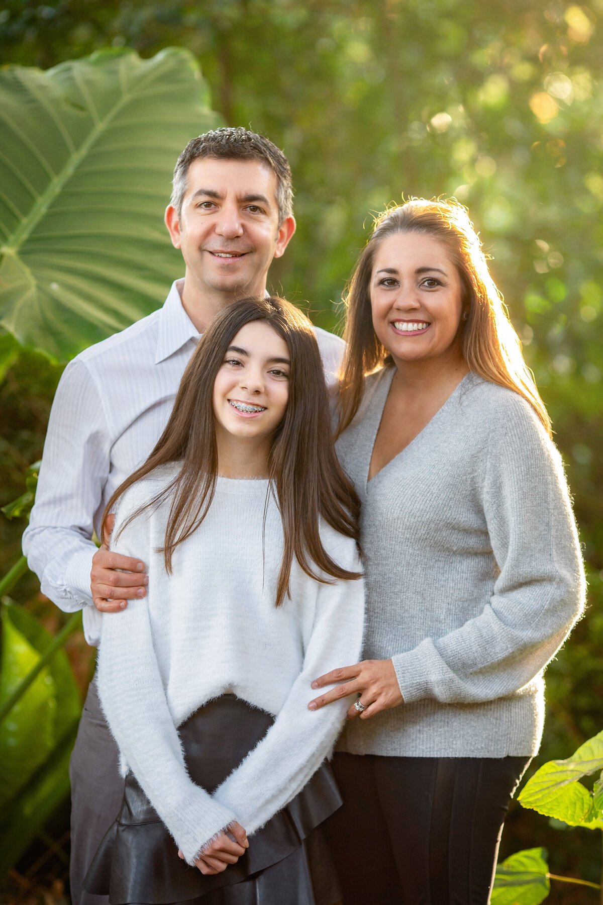 Family of 3, backlit in a jungle-like setting.  Daughter and mom have long brown hair.  They are wearing sweaters and black pants.  Dad is wearing a gray shirt.