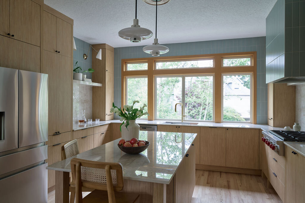 Kitchen with white oak cabinets, and slim simple black hardware. Taj mahal quartzite countertops that run up the backsplash to a quartz shelf with cookbooks and plants on it. Green subway tile backsplash above the shelf to the ceiling. Two sconces above the shelf that are modern, white and gold.