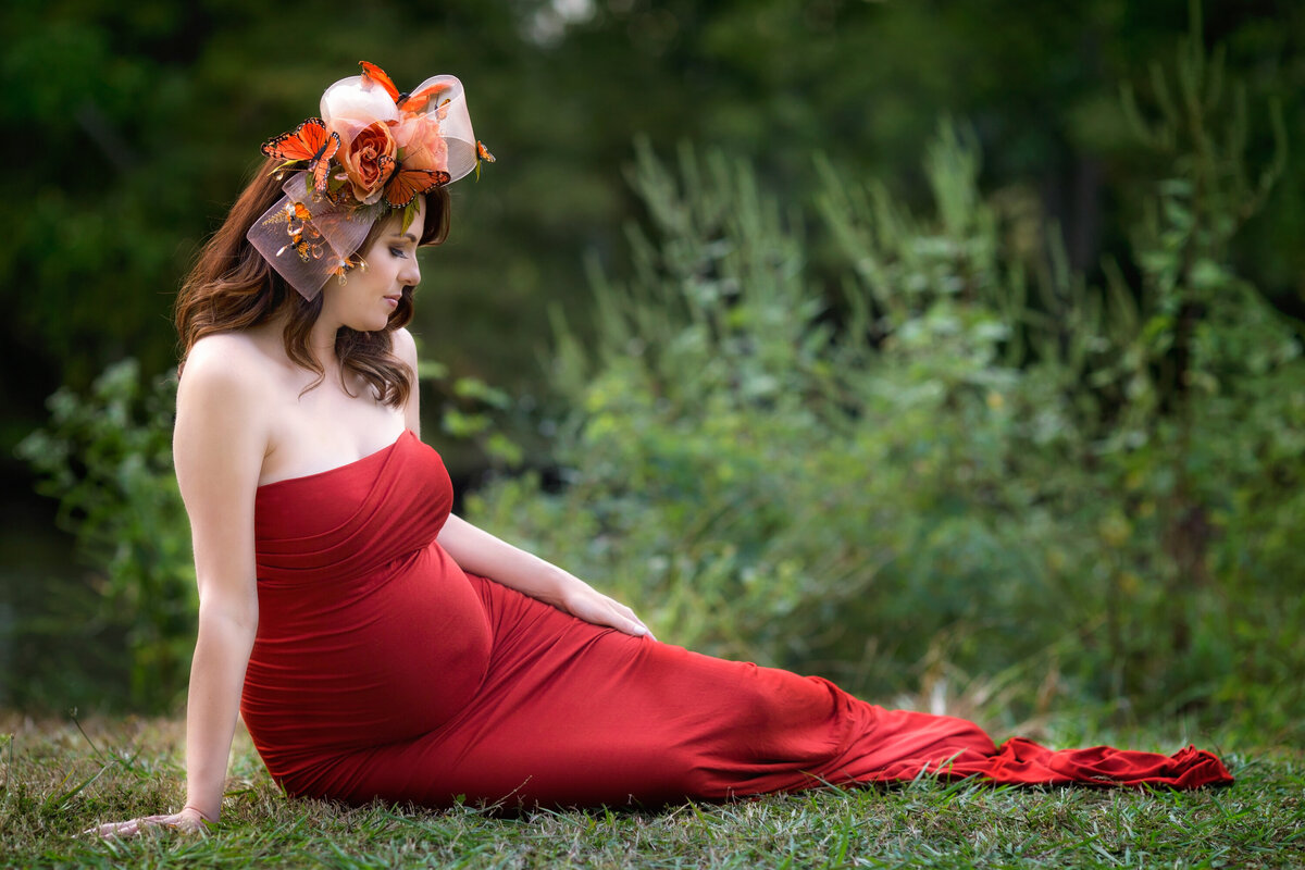 Pregnant woman in an orange Sew Trendy dress sits on the banks of the bayou in Audubon Park.  She has a custom butterfly headpiece from Miele Moda.  Her pregnant belly shows and her belly button pops out.  She is looking at her belly.