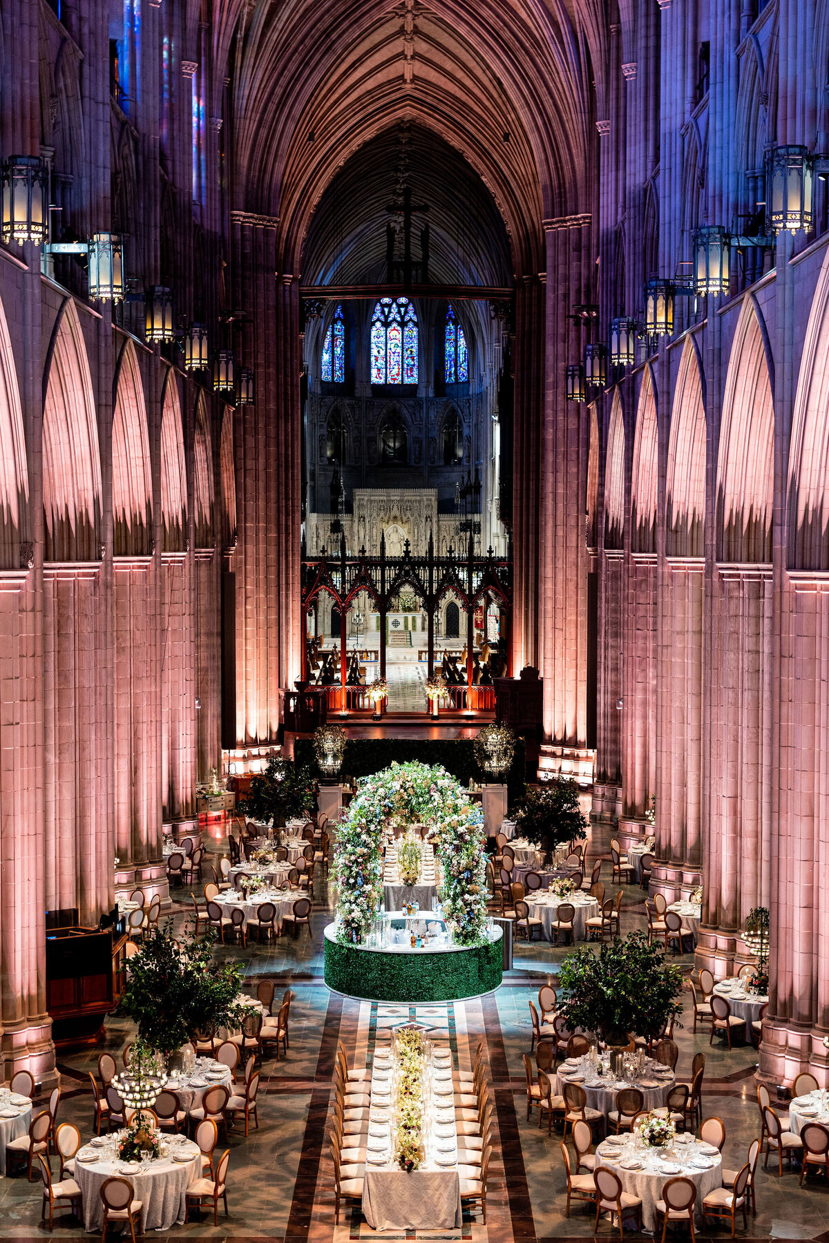 A grand, gothic-style cathedral with high vaulted ceilings hosts an elegant event. Round tables with white tablecloths and greenery surround a central floral arrangement. Soft, warm lighting and ornate stained glass windows enhance the majestic atmosphere.