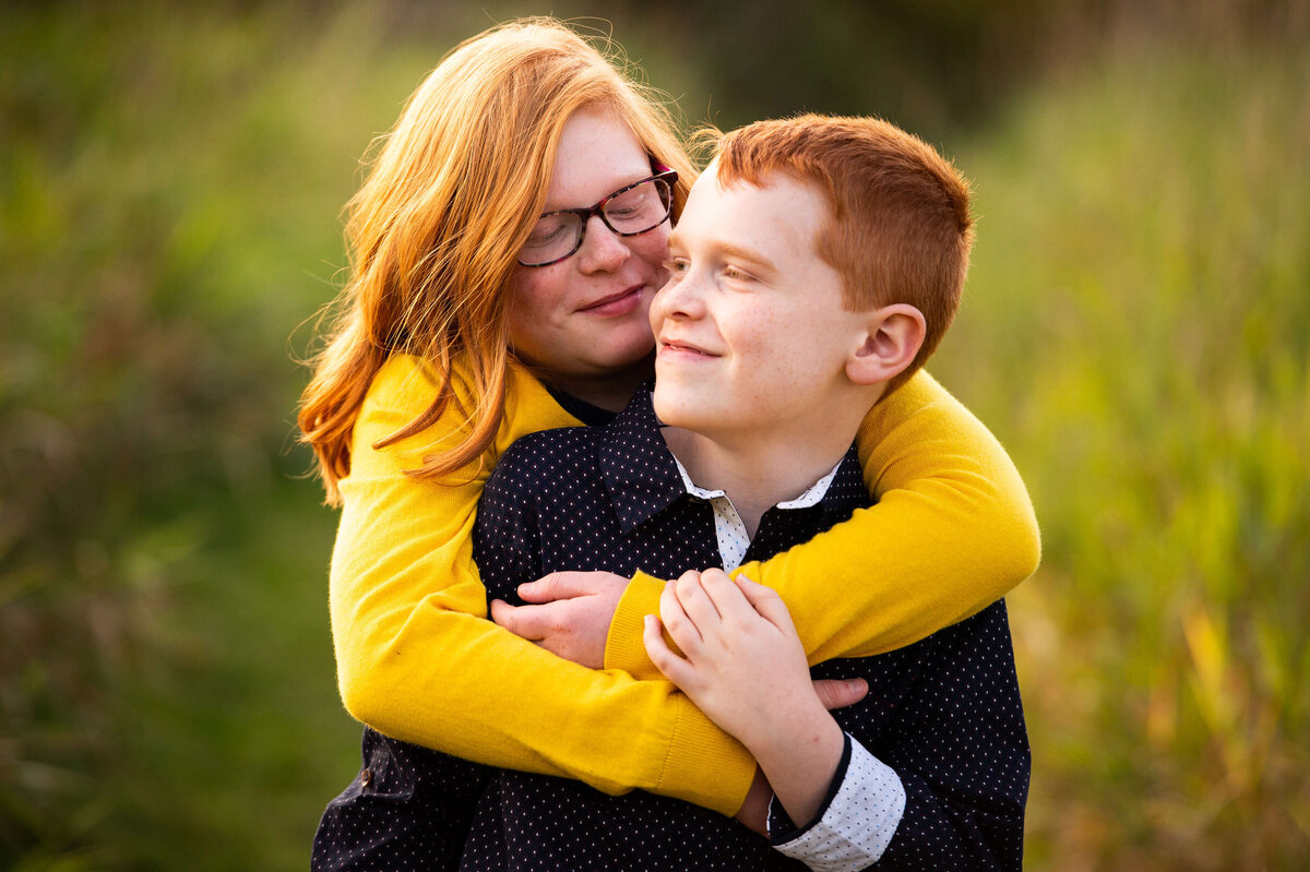 family photography in Ottawa showing an older sister hugging her younger brother in a grassy field at sunset