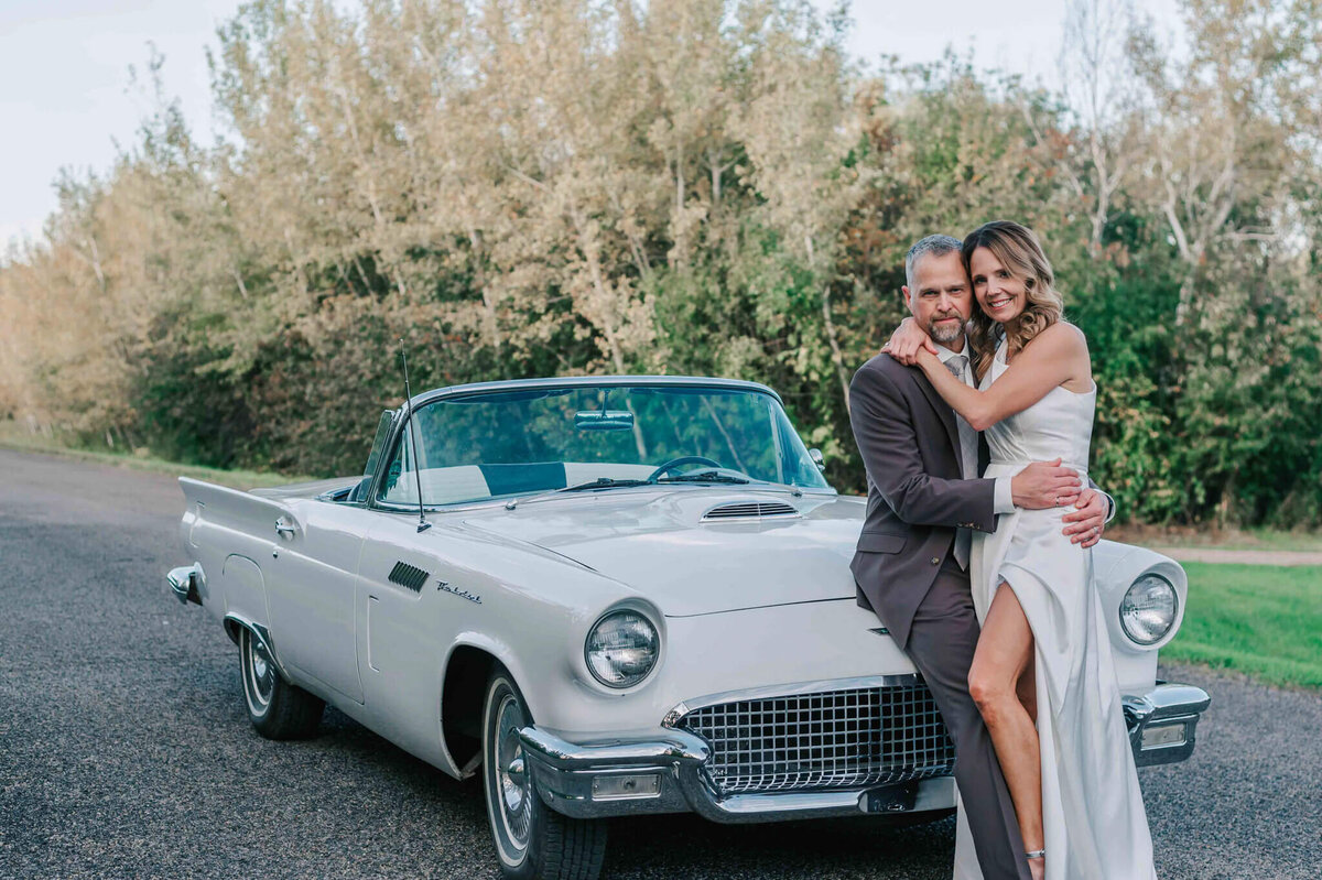 Bride and groom sitting on the hood of a car holding each other.