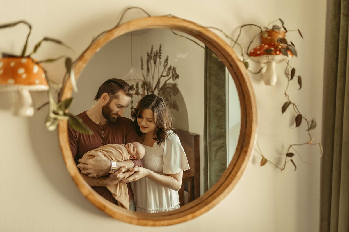 Reflection of a mother , father and baby in the mirror. The nirror has a wooden frame and there are mushroom shaped lamps on both sides.