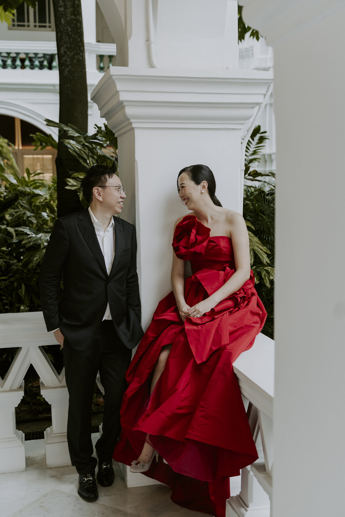 a bride in a red dress and a man in a black suit smile at each other on the balcony