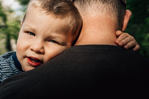 A toddler boy looking back over his father's shoulder.