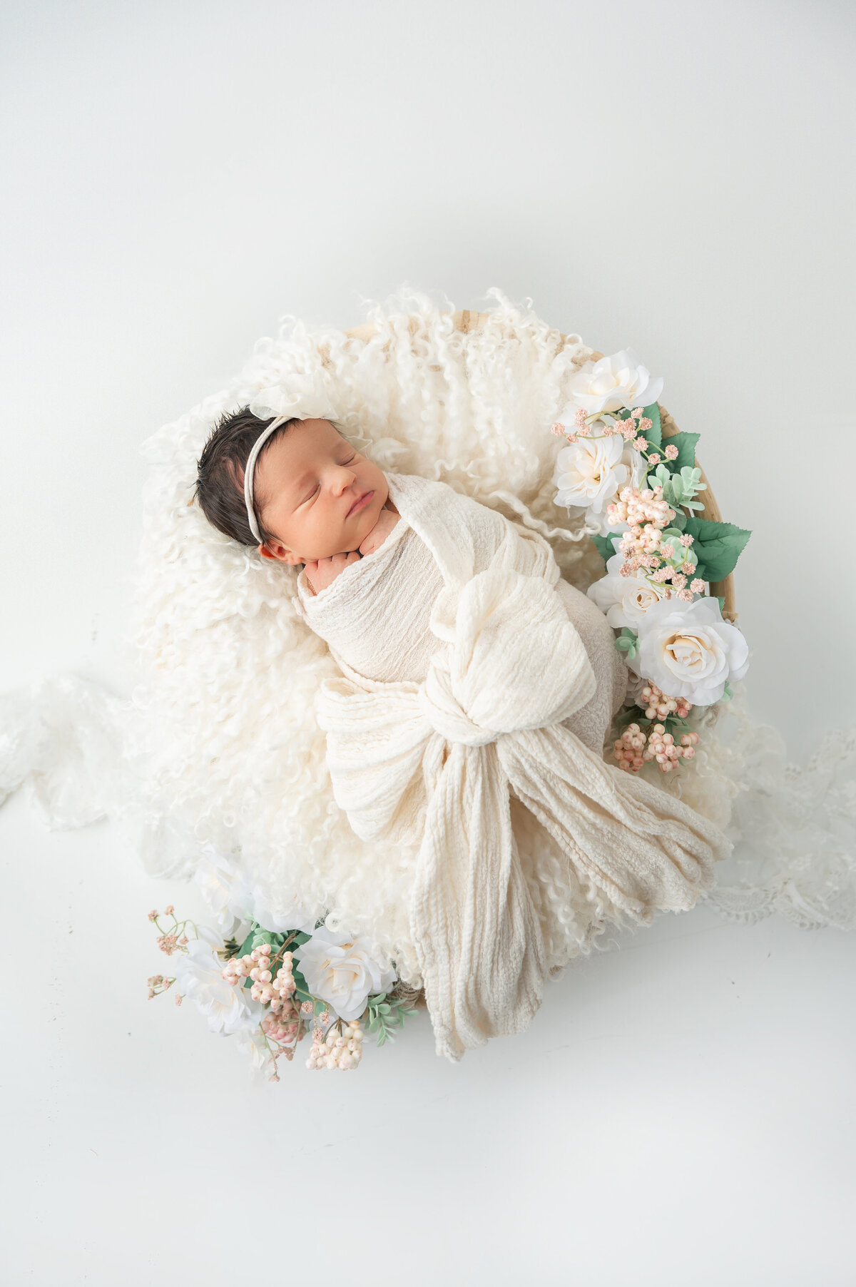 Baby swaddled in off-white wrap with a bow, resting on flokati surrounded by coordinating colored roses.
