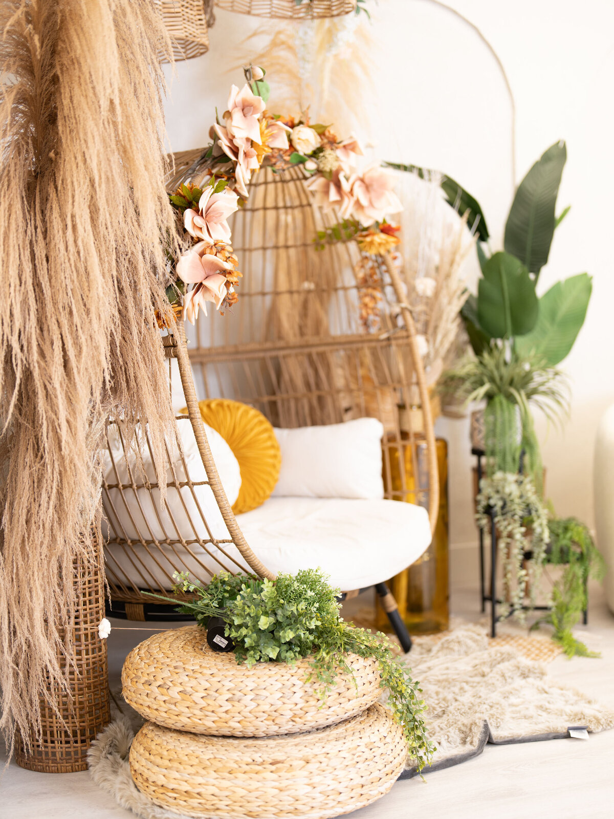 Cozy bohemian nook with a wicker chair and white cushion, adorned with pink flowers and pampas grass. Vibrant greenery in baskets on a stylish rug.