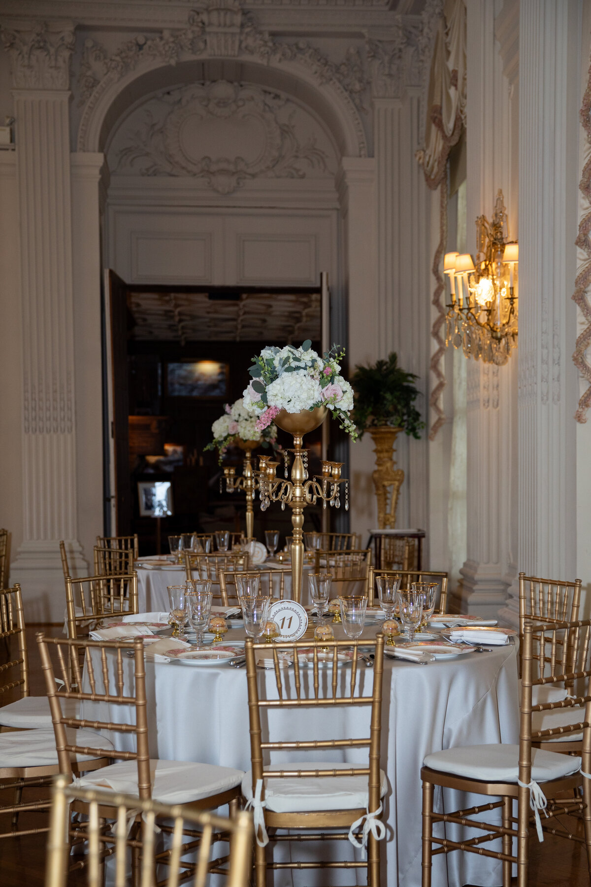 Wedding Photographers in RI | Kelsey Sheehan Photography Timeless Rhode Island Weddings | Elegant dining room with a round table set for an event, featuring white linens, golden chairs, a floral centerpiece, and soft wall lighting, conveying a formal tone.