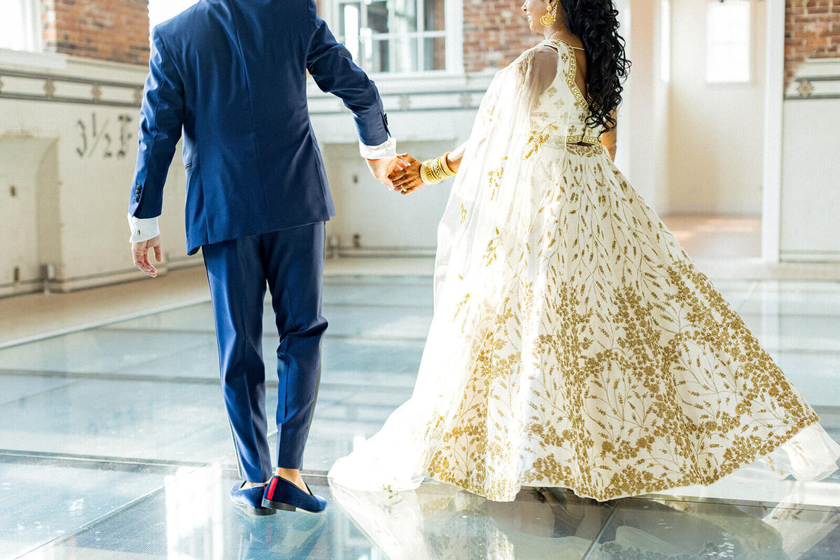  Indian bride and groom holding hands while walking across the glass floor at 701 Whaley during their vibrant three-day Indian wedding weekend in South Carolina. The groom wears a navy blue suit, and the bride stuns in a white and gold embroidered lehenga, blending cultural elegance with the venue’s modern charm. A stunning moment captured in luxury South Carolina wedding photography.