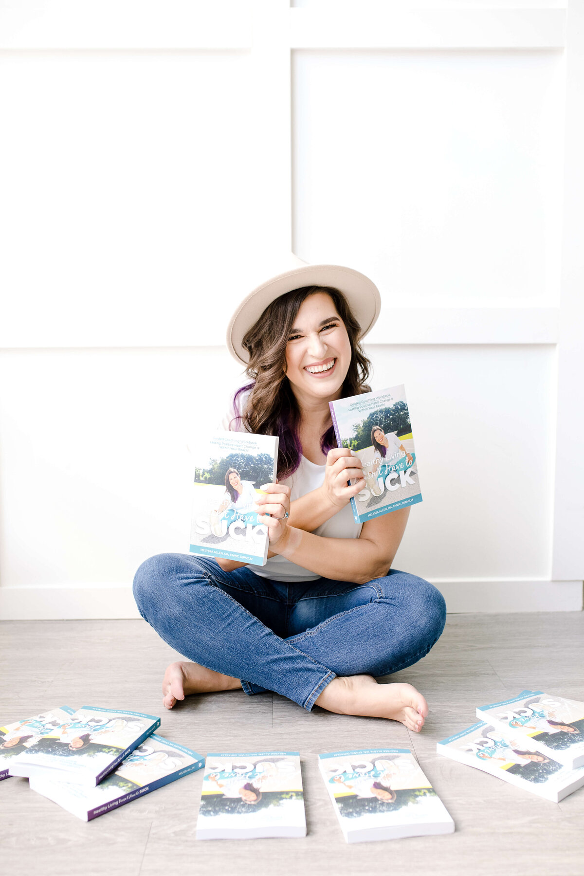Brand photo shoot ideas for an author with woman sitting behind a pile of books while holding two of the books up to show her new book as she smiles while wearing a wide brimmed hat