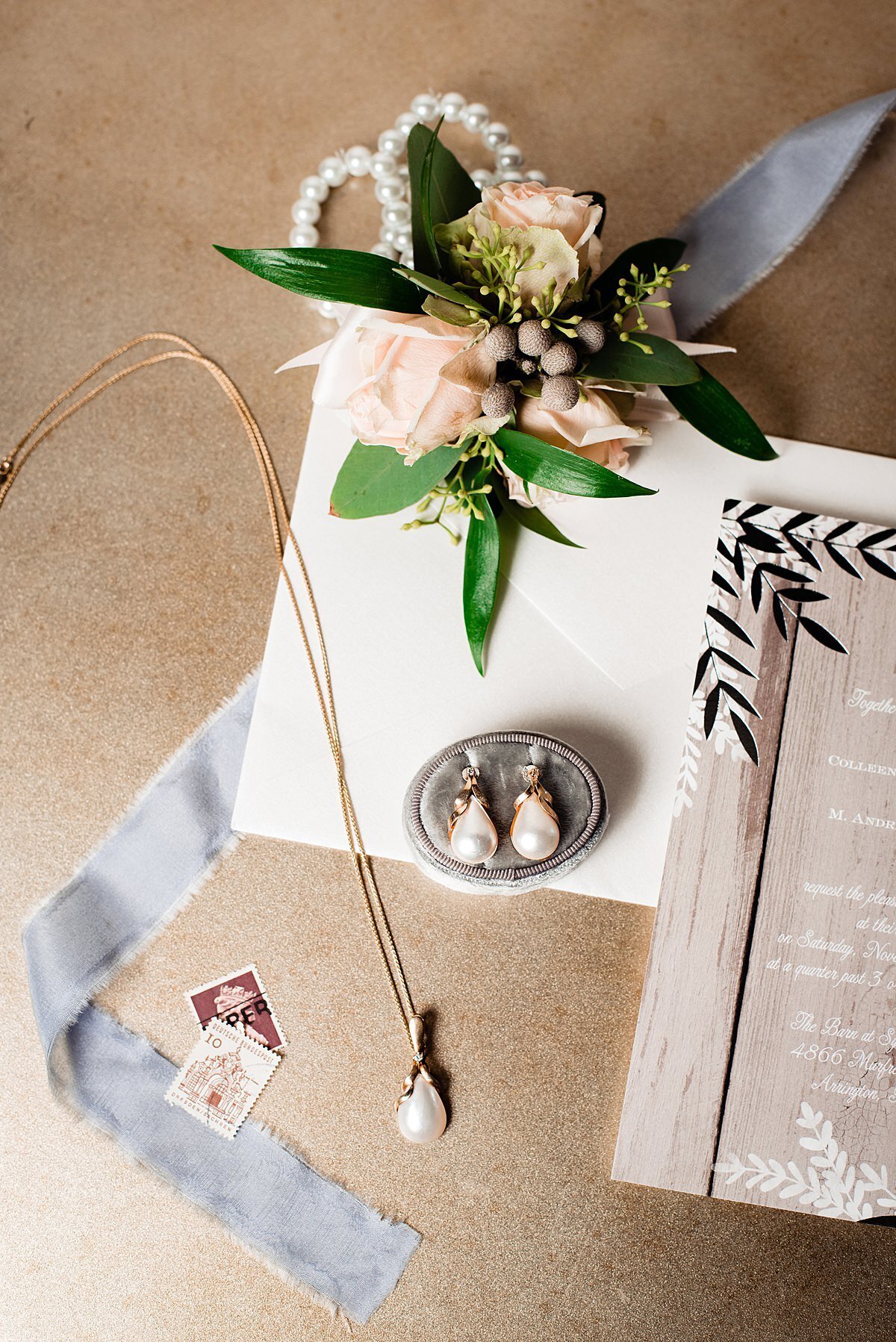 The edge of the wedding invitation and a blue raw silk ribbon with some vintage stamps accent the bride's baroque drop pearl necklace on a gold chain as well as her matching baroque drop pearl earrings set in gold next to her wedding garter and a peach dahlia flower