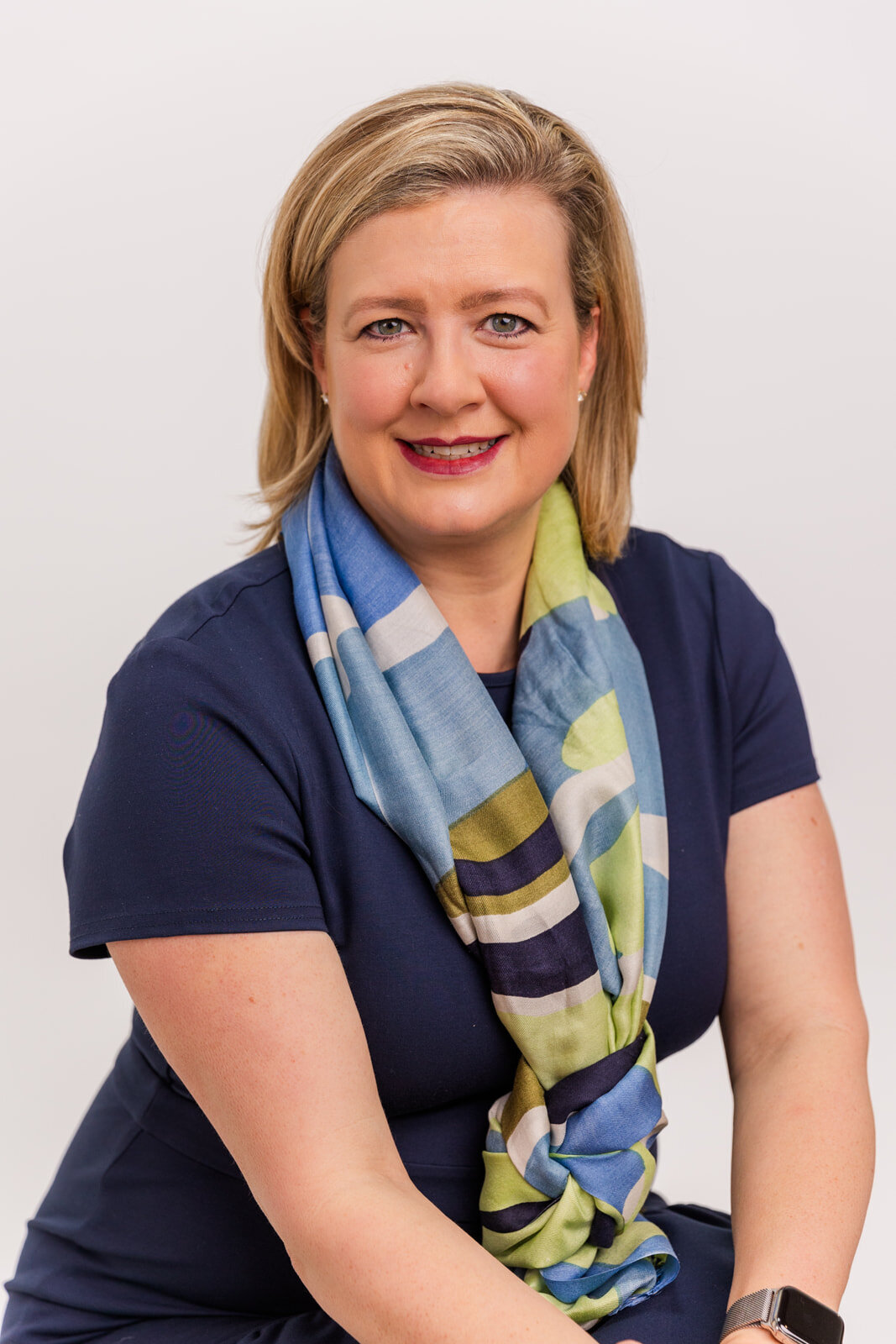 Corporate portrait of woman wearing a navy blue dress and statement scarf around her neck sitting on a stool in front of a white background in Buckhead studio