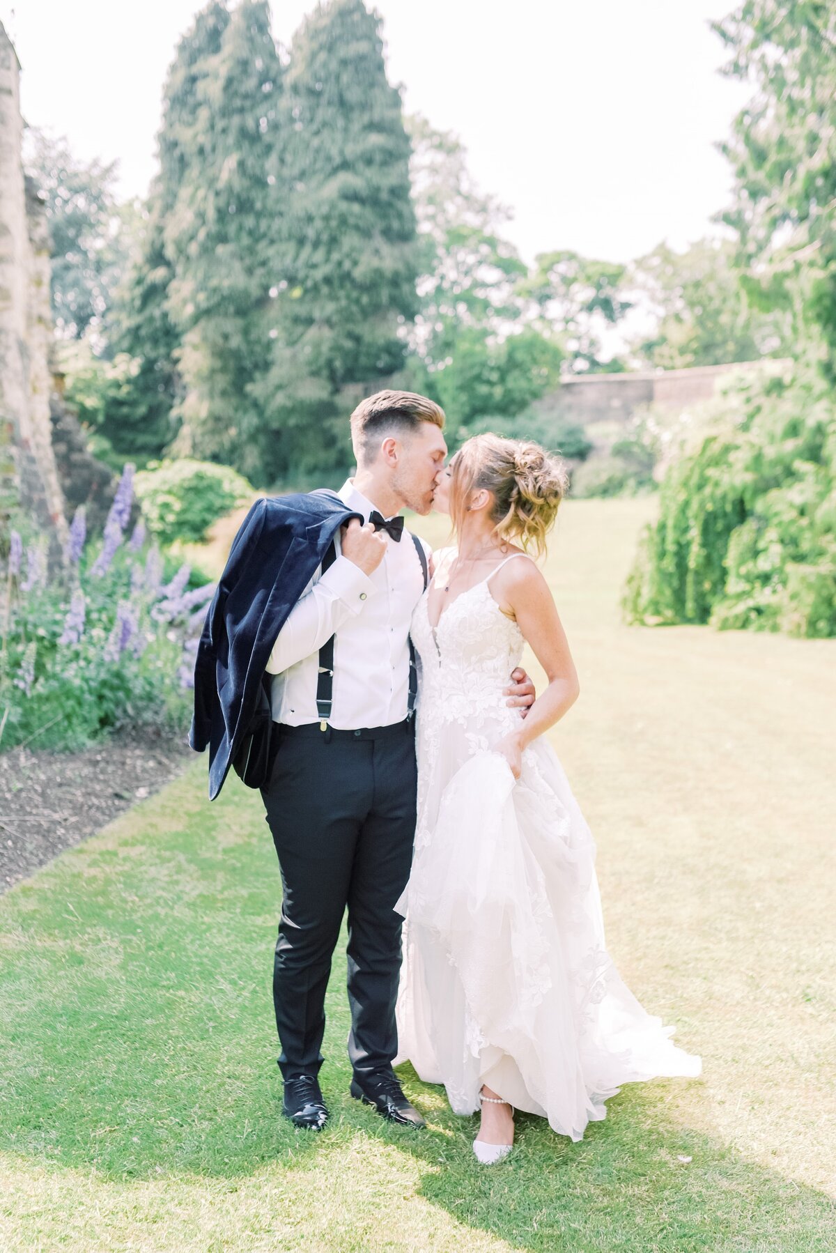 Light and airy style image of a bride and groom walking together, they have stopped for a kiss. The groom is in a black tuxedo and the bride is wearing a thin strap aline lace gown