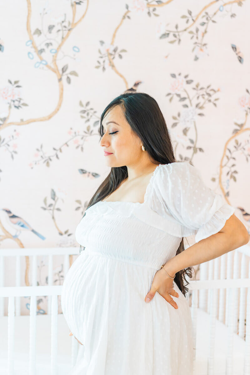 A pregnant woman dressed in a white dress with long dark hair stands in front of a crib in a pink nursery. 