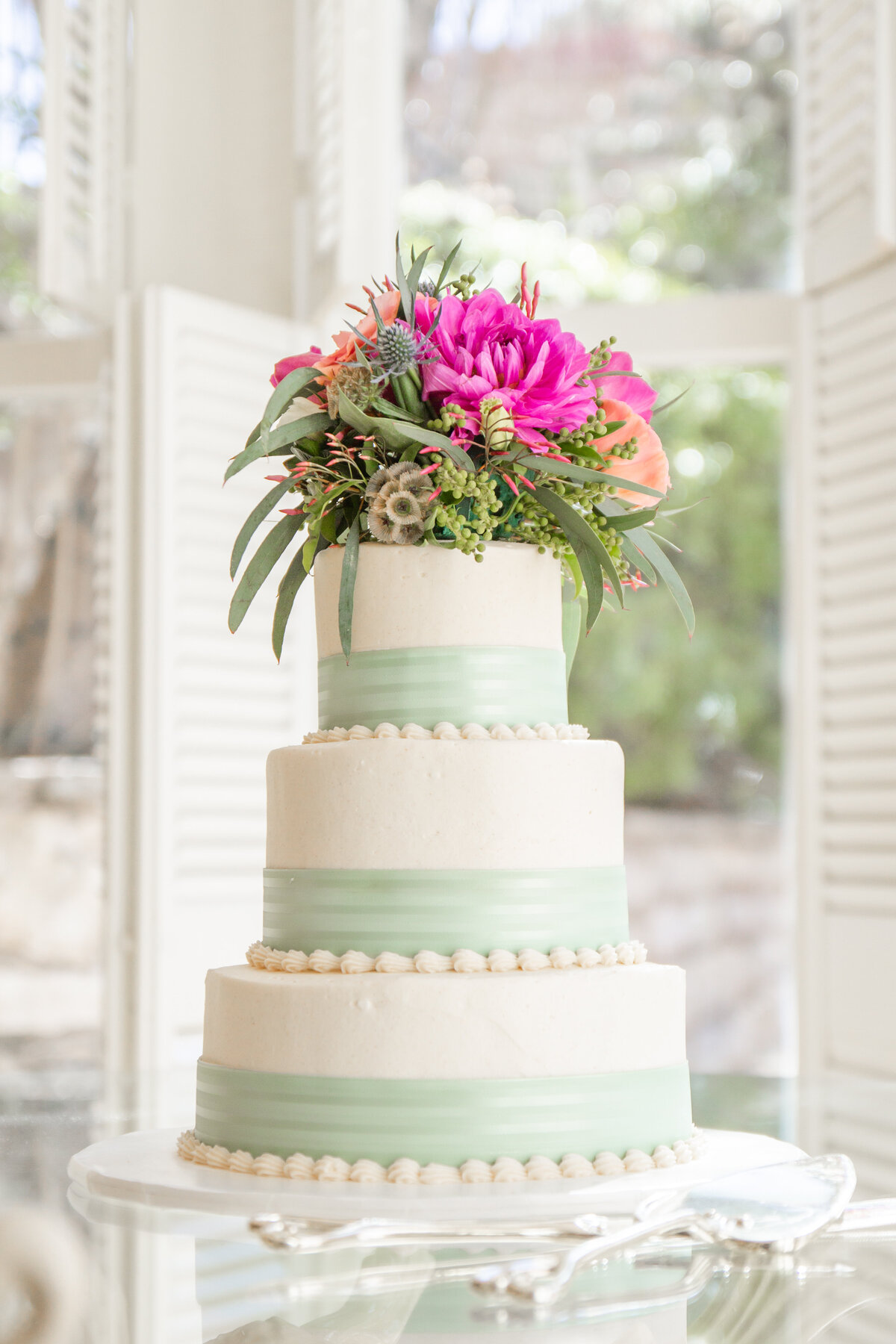 3 layer wedding cake with mint green ribbon and pink flowers on table with shutters at Chateau Bellevue in Austin Texas