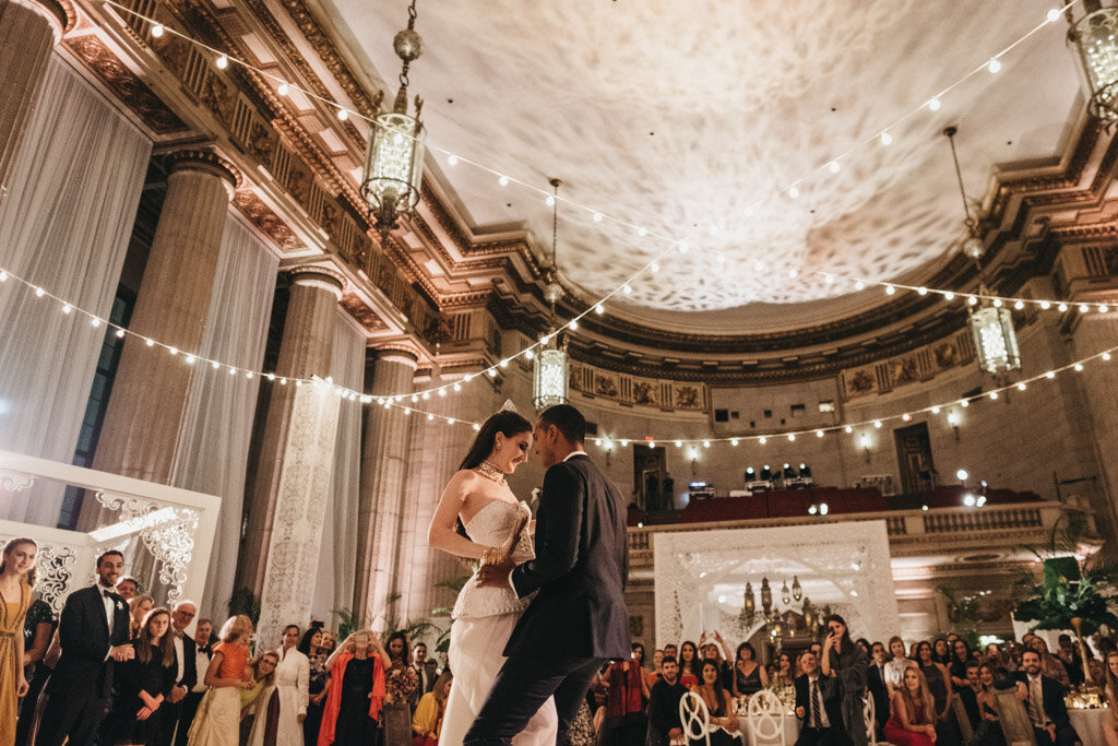 This is where the photo was taken at Washington D.C., The Mellon Auditorium, one of the best luxury DC venues with European flair, beautifully captured by photographer Sarah Bradshaw. A bride and groom share their first dance in a grand, ornate hall with high ceilings, columns, and hanging lanterns. String lights create a warm atmosphere, while guests surround them, watching and smiling. The hall's decor exudes elegance and historic charm.