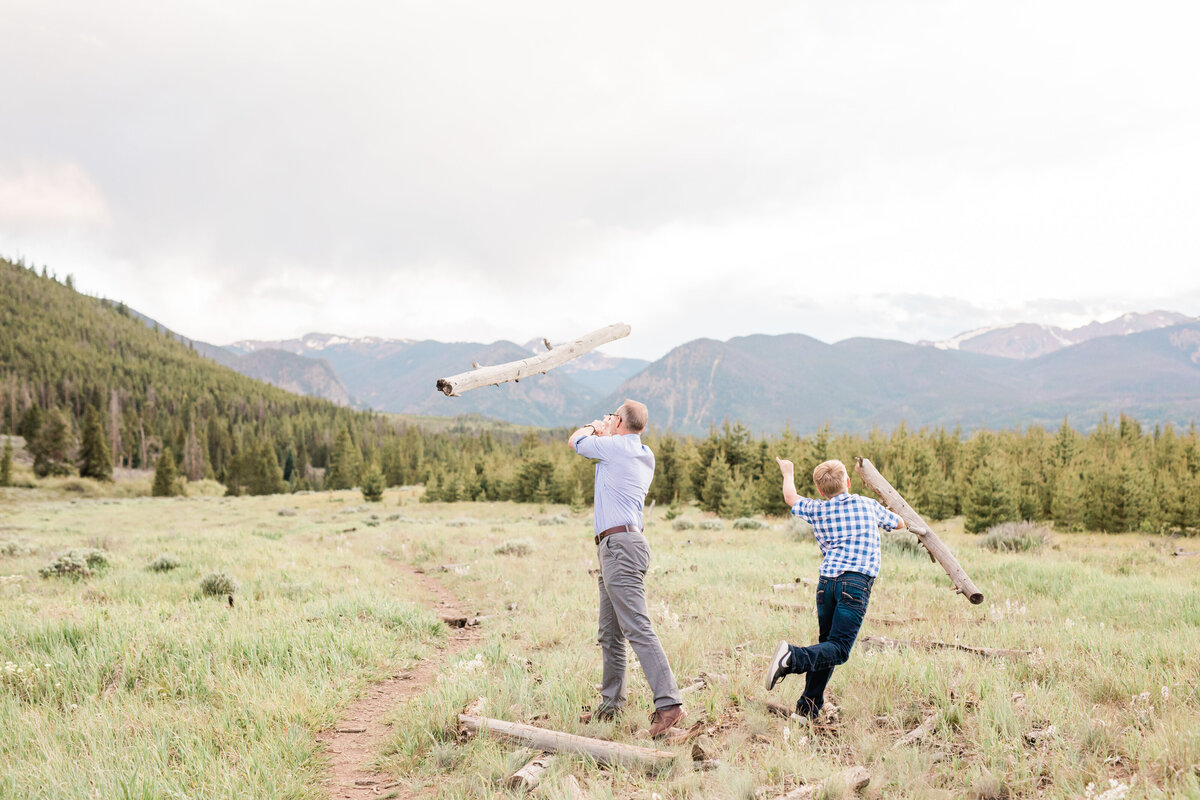 This father and son are seen in a large mountain meadow. We can see them from the back as they have a log throwing contest to see who can throw the furthers. The logs are mid-air in this photo