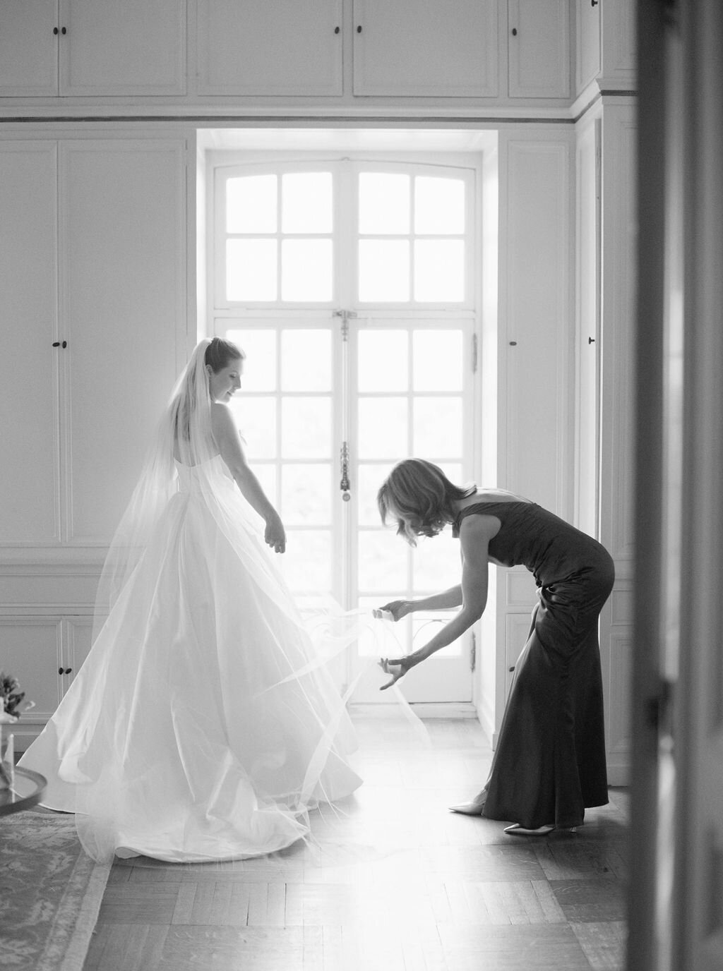 Rhode Island Wedding Photographers | A bride in a flowing gown stands by a bright window as another woman adjusts the hem. 