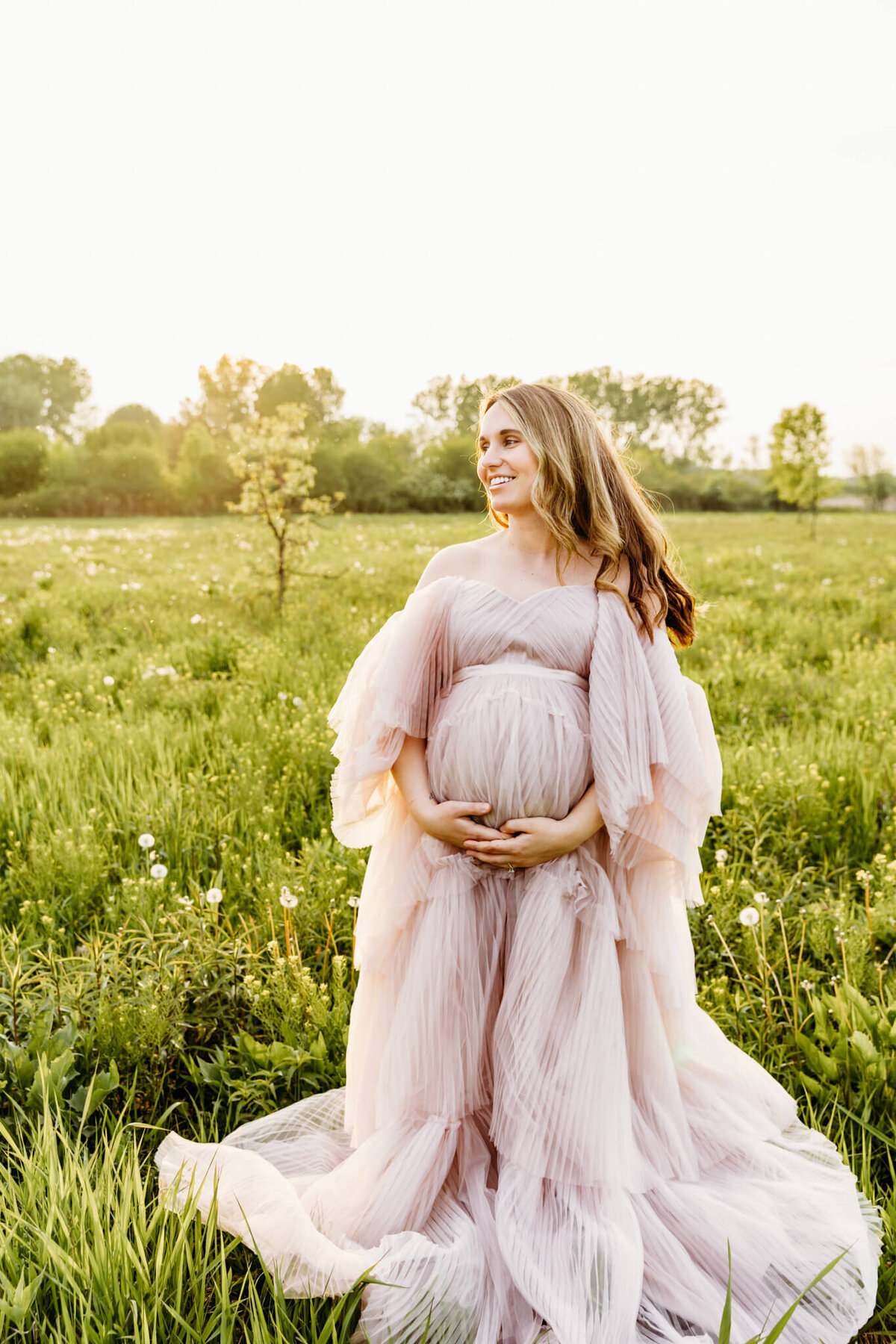 beautiful expecting mother in a light pink gown holding her baby bump and looking over her shoulder in a field at sunset near Green Bay