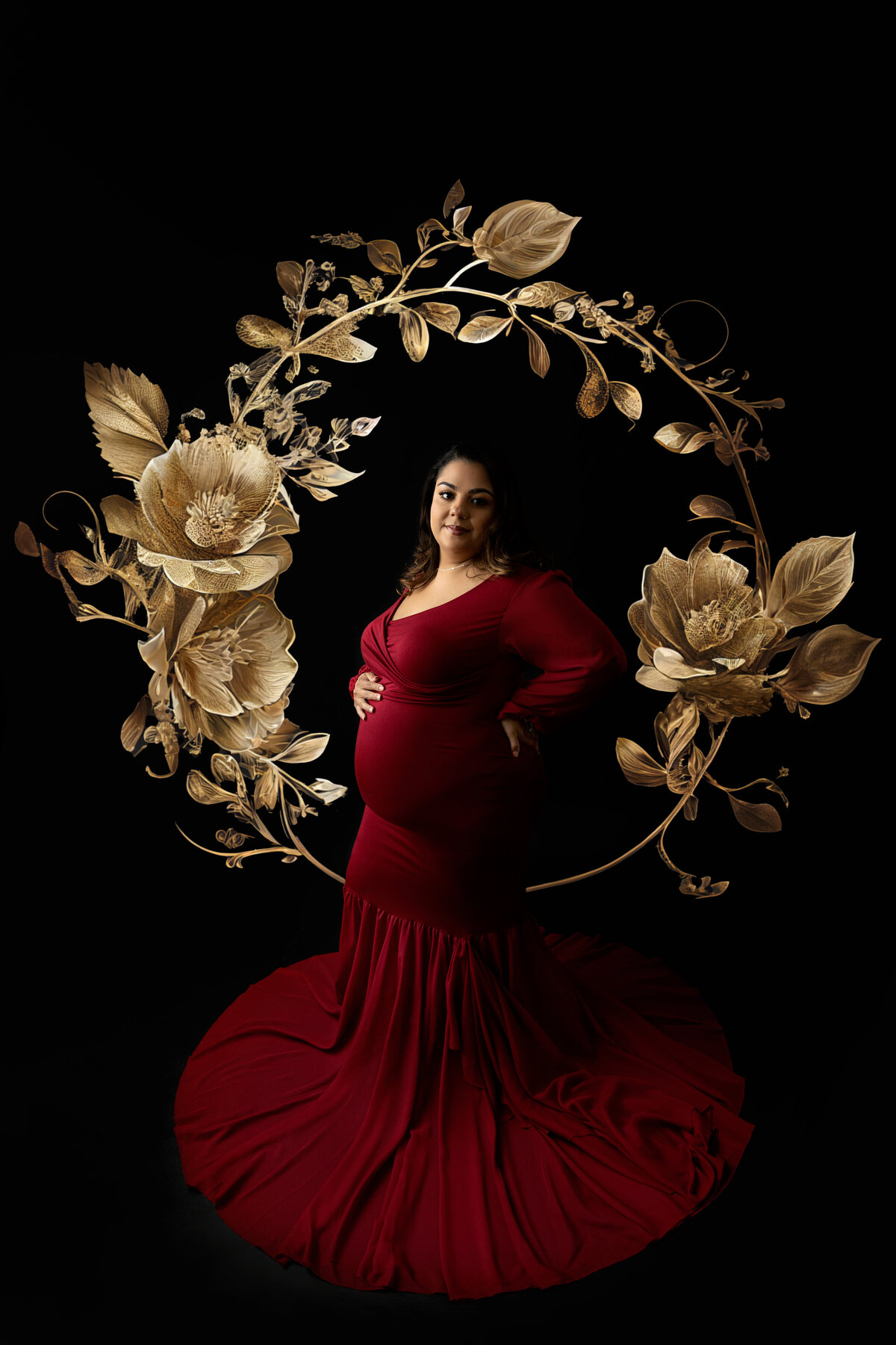 A pregnant woman in a red maternity gown stands in a studio with a gold halo in the background