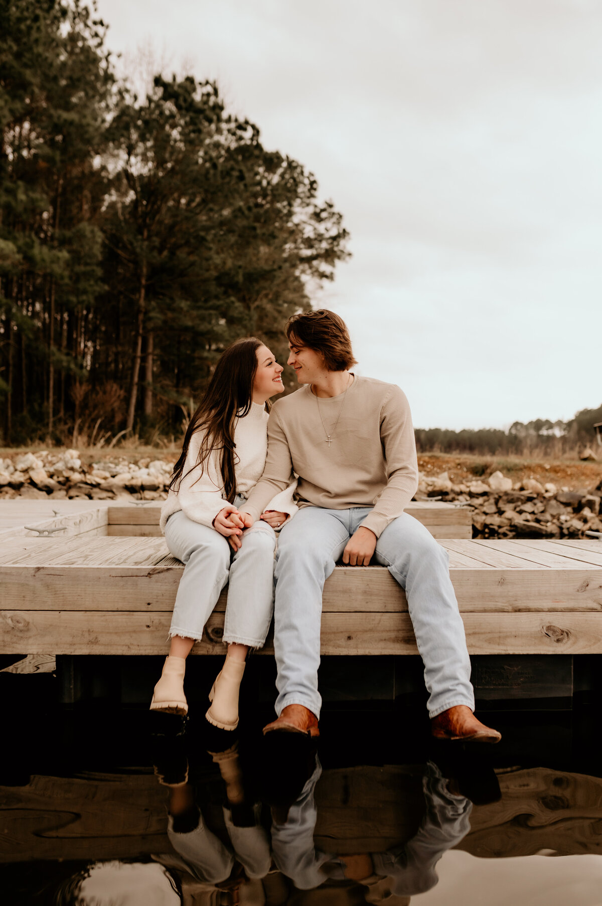 Little rock engagement photographer photographs man and woman sitting together ona dock at a lake while holding hands and leaning into each other and smiling