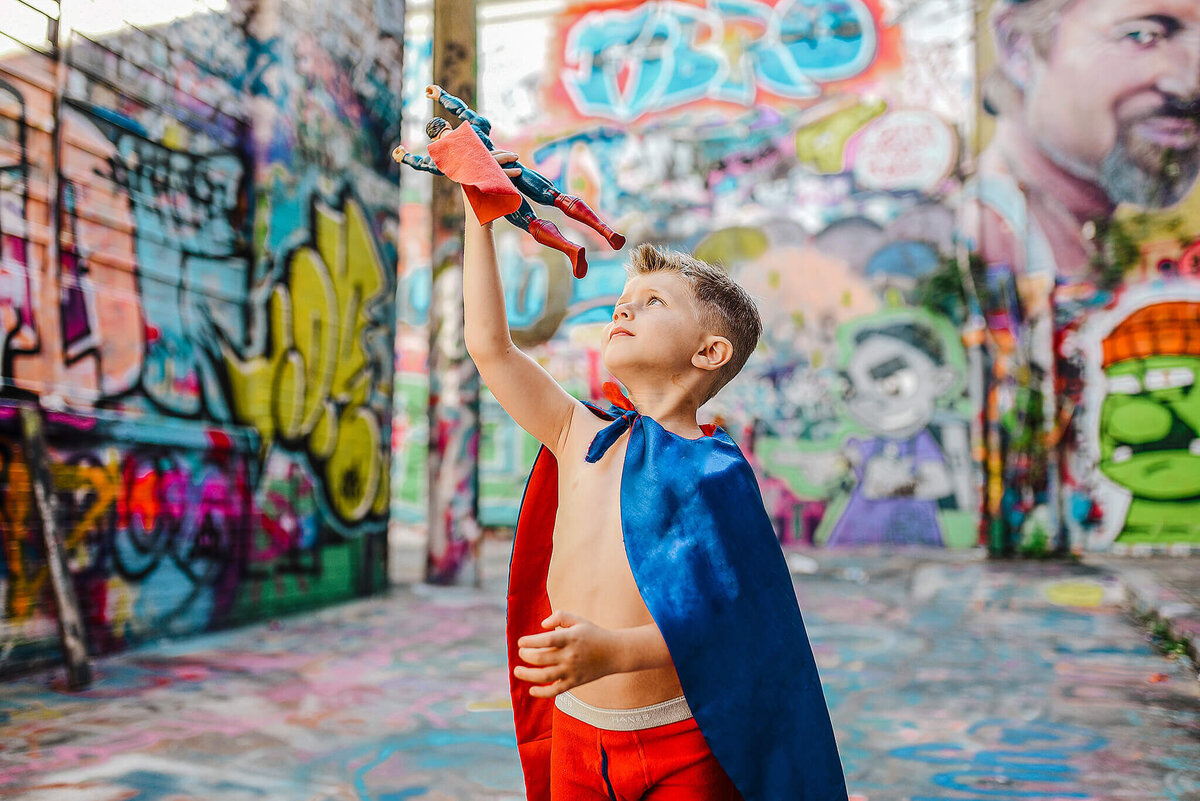Little boy in red underwear and blue superhero cape playing with a superman action figure in graffiti alley near MICA in Baltimore Maryland
