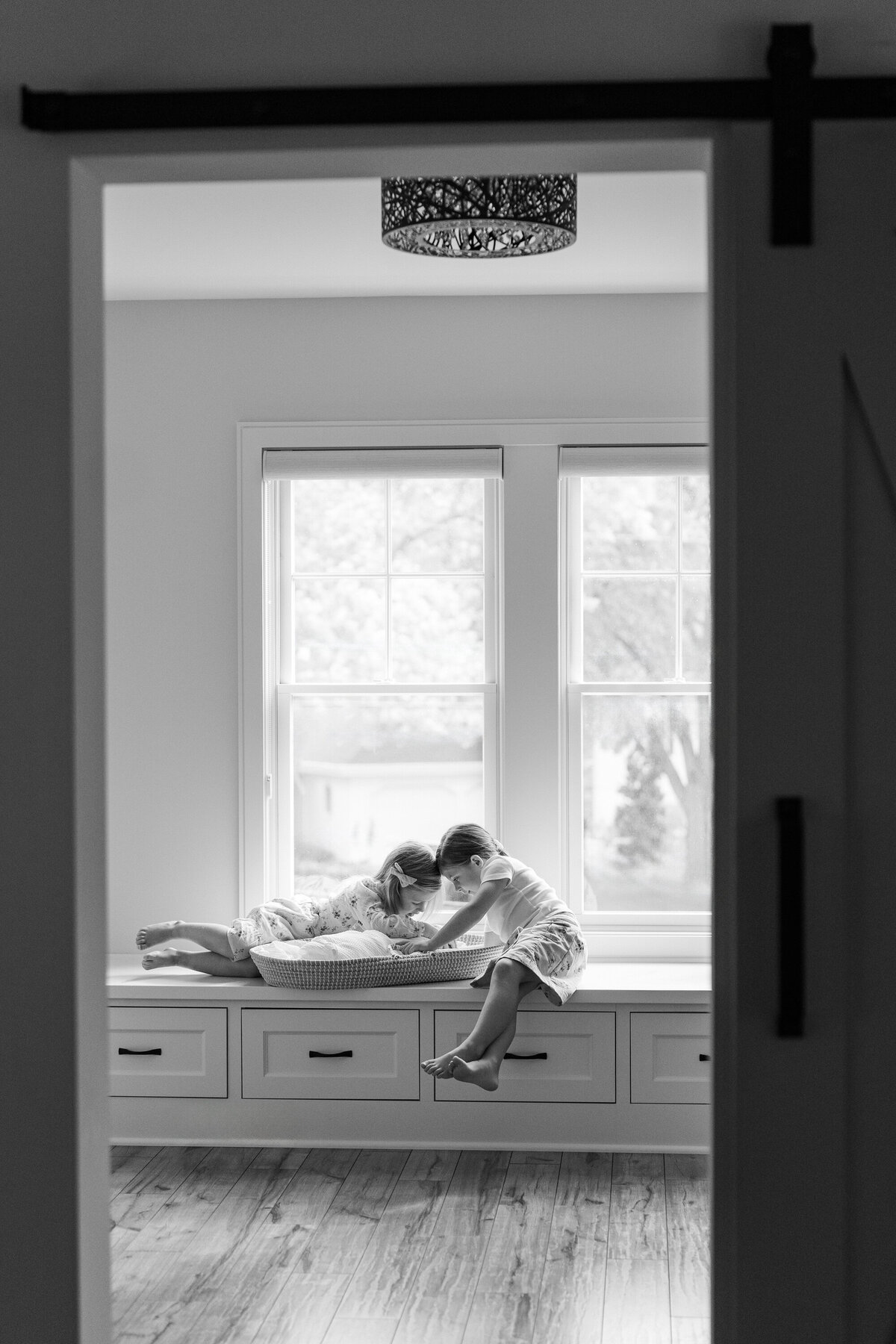 Two older siblings sitting in a window, happily gazing at their newborn sibling who is nestled in a basket