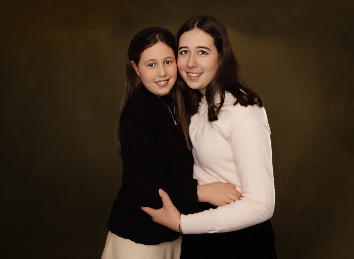 Sisters pose for a family studio photoshoot in brooklyn, ny. The sisters, both teens are standing facing each other with their cheeks touching and smiling at the camrea. Captured by premier Brooklyn NY family photographer Chaya Bornstein Photography.