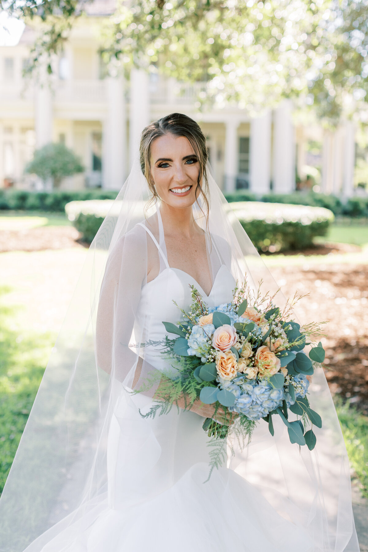 A bride holds her peach and blue bouquet while smiling at the camera.