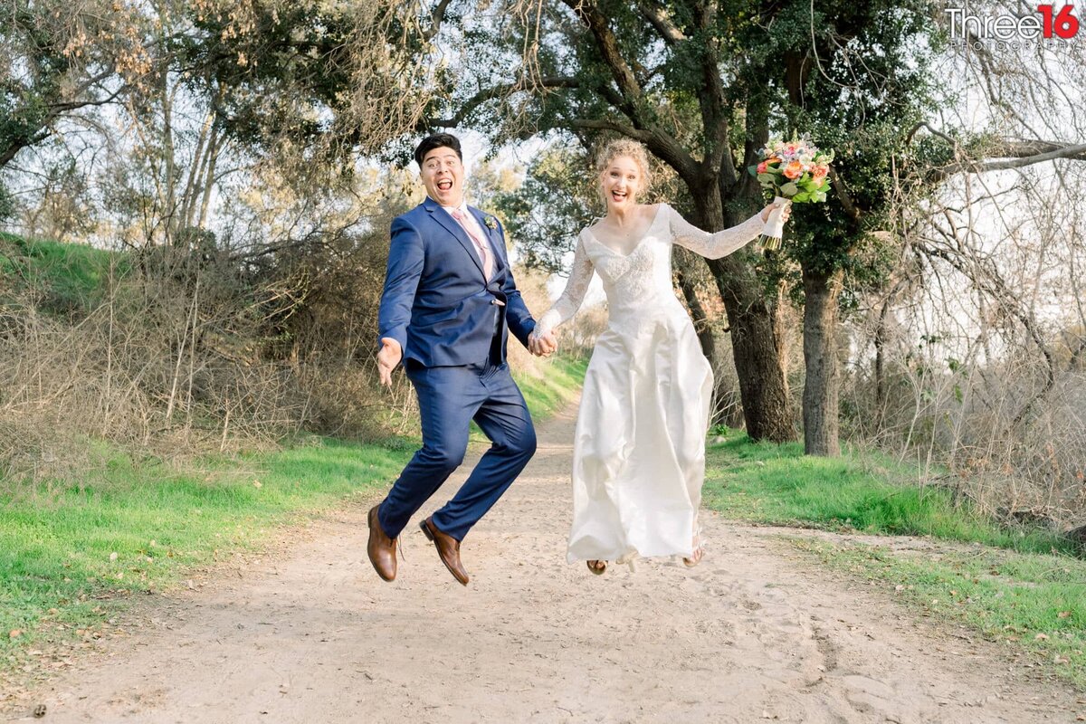 Bride and Groom jump for joy during their wedding photo session as they walk along a dirt trail