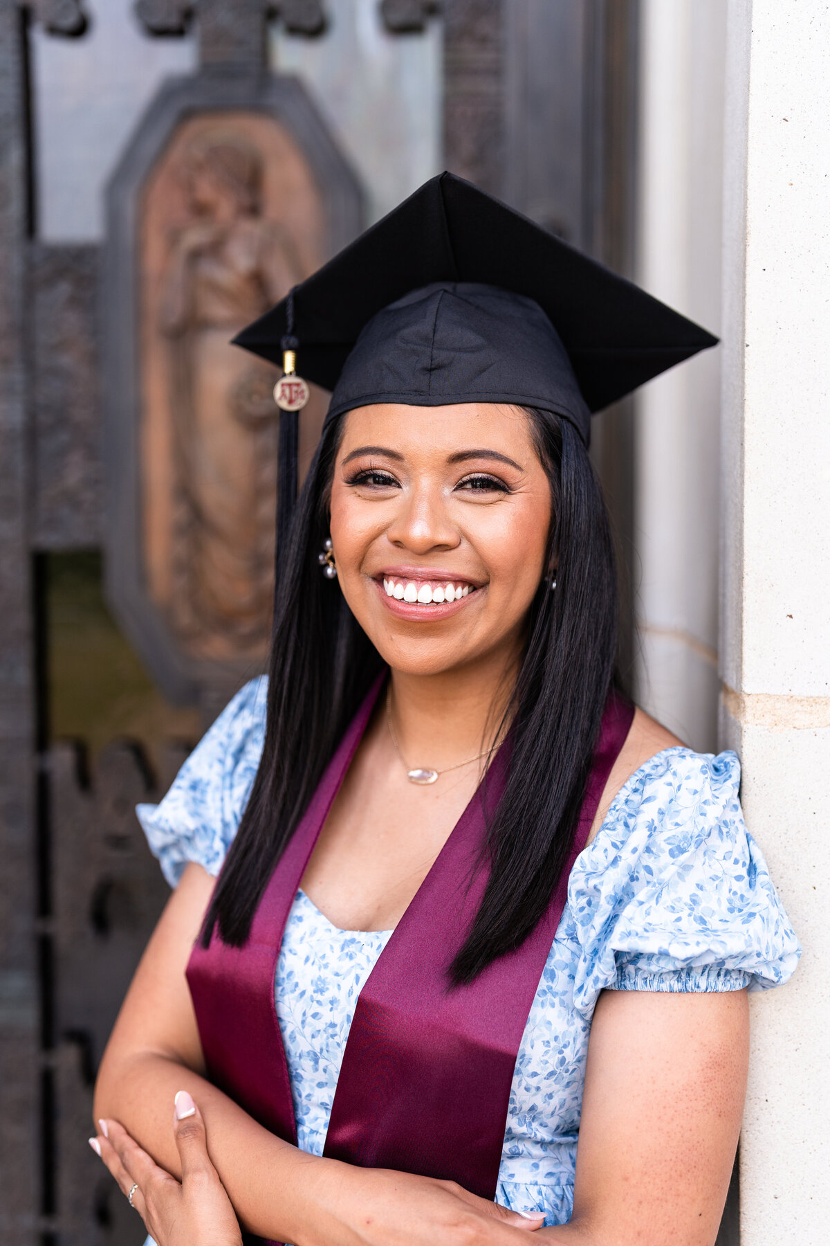 Texas A&M senior girl crossing arms and smiling while wearing blue dress, maroon stole and grad cap while leaning on door of Administration Building