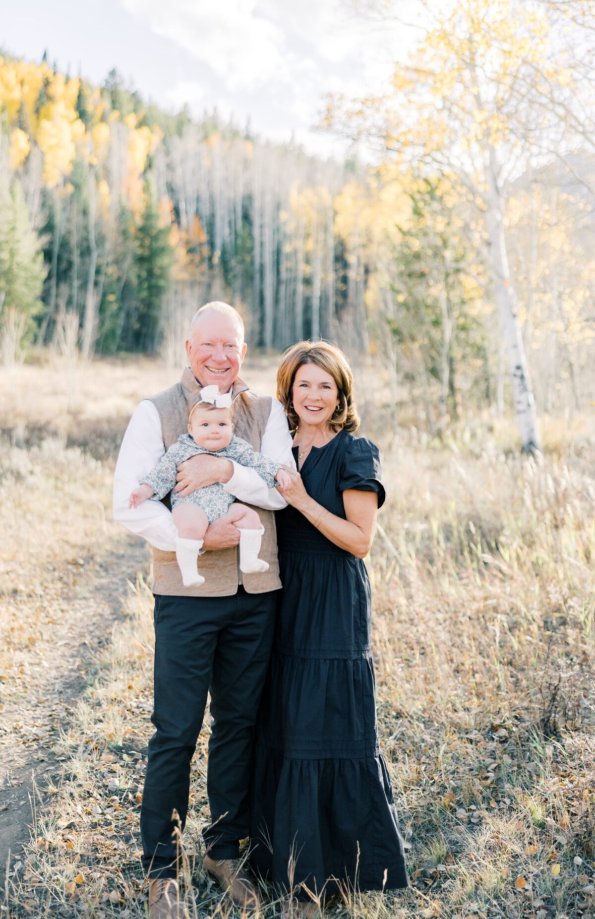 This is a portrait of a grandma and grandpa while they hold their young grandchild. They are all looking at the camera, surrounded by the fall aspen leaves in Breckenridge, Colorado. 