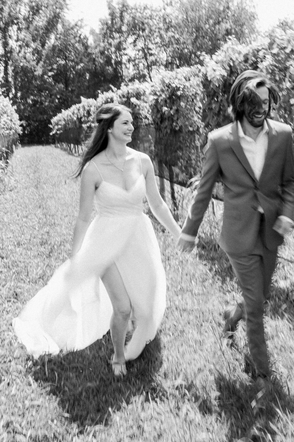 A black and white photo of a bride in a flowing dress and a groom in a suit, holding hands and walking through a grassy area with a sense of joy.