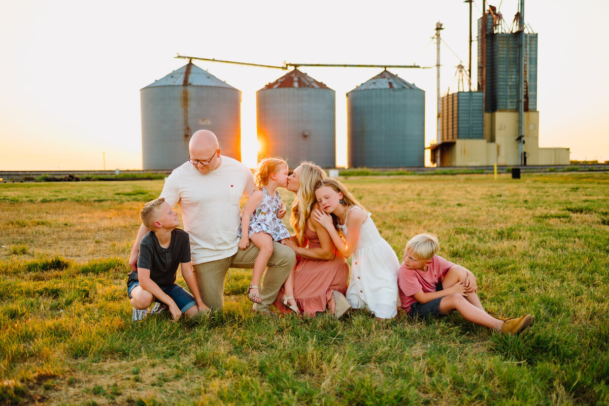 Family photo sessions in Albuquerque captured in front of a big farm with a vintage charm. The family is sitting on green grass, dressed in vintage outfits. The parents and their four kids are all smiling and appear joyful, embodying the essence of a warm