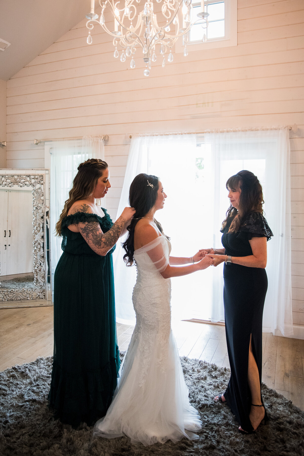 A bride stands with two bridesmaids. One holds her hands and the another puts on her necklace.