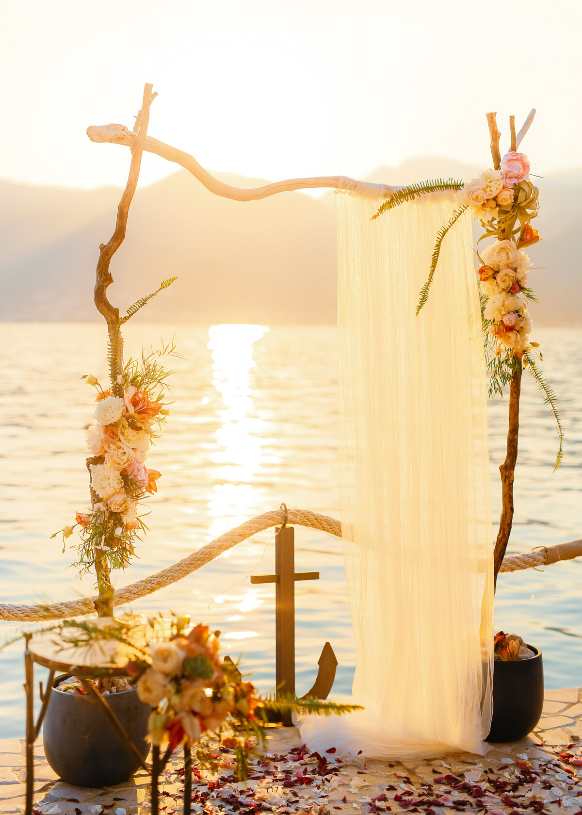 A ceremony arch is decorated with flowers and a drape against the ocean at sunset in Greece.