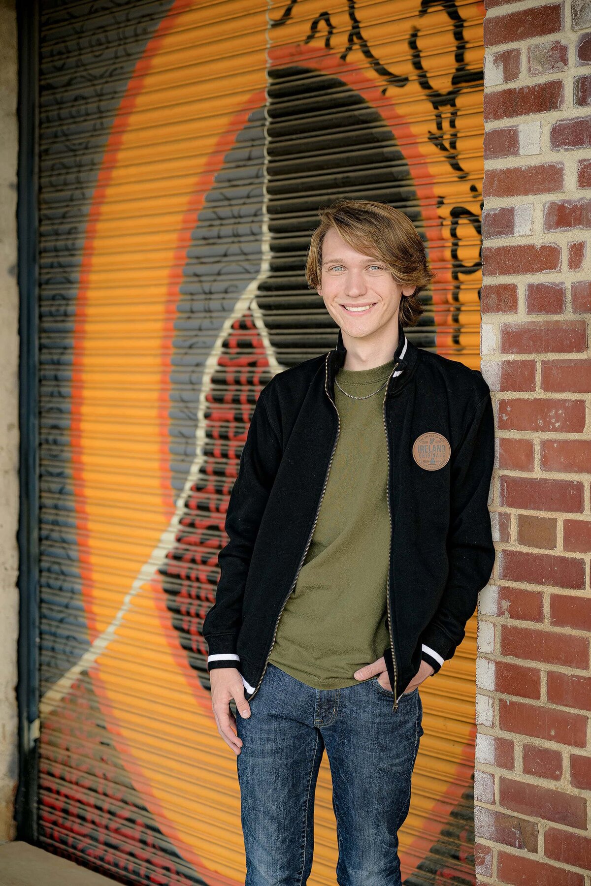 Senior in high school stands wearing a green shirt and jacket and his hands in his pockets at an urban venue